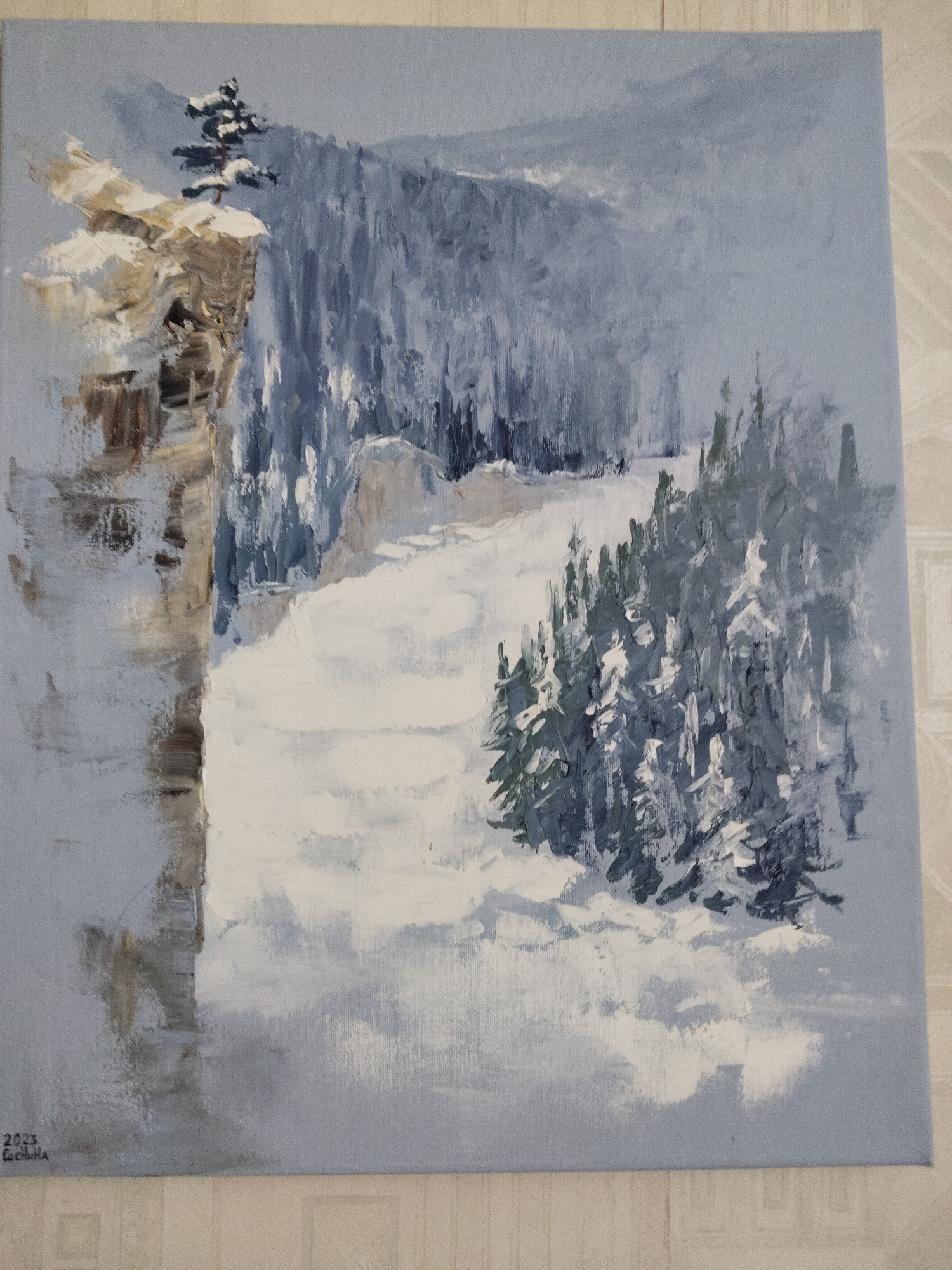 A painting depicts a snowy, mountainous landscape. A large, tan-colored rock formation with snow on top sits on the left side of the painting, with a solitary pine tree growing from its peak. A forest of evergreen trees covered in snow fills the middle ground, leading to a snow-covered valley or clearing. In the background, a mountain range fades into the distance, partially obscured by mist or snow. The overall color palette is dominated by shades of white, grey, and blue, suggesting a cold, wintry environment. The painting is signed "2023 Cecilia" in the bottom left corner.