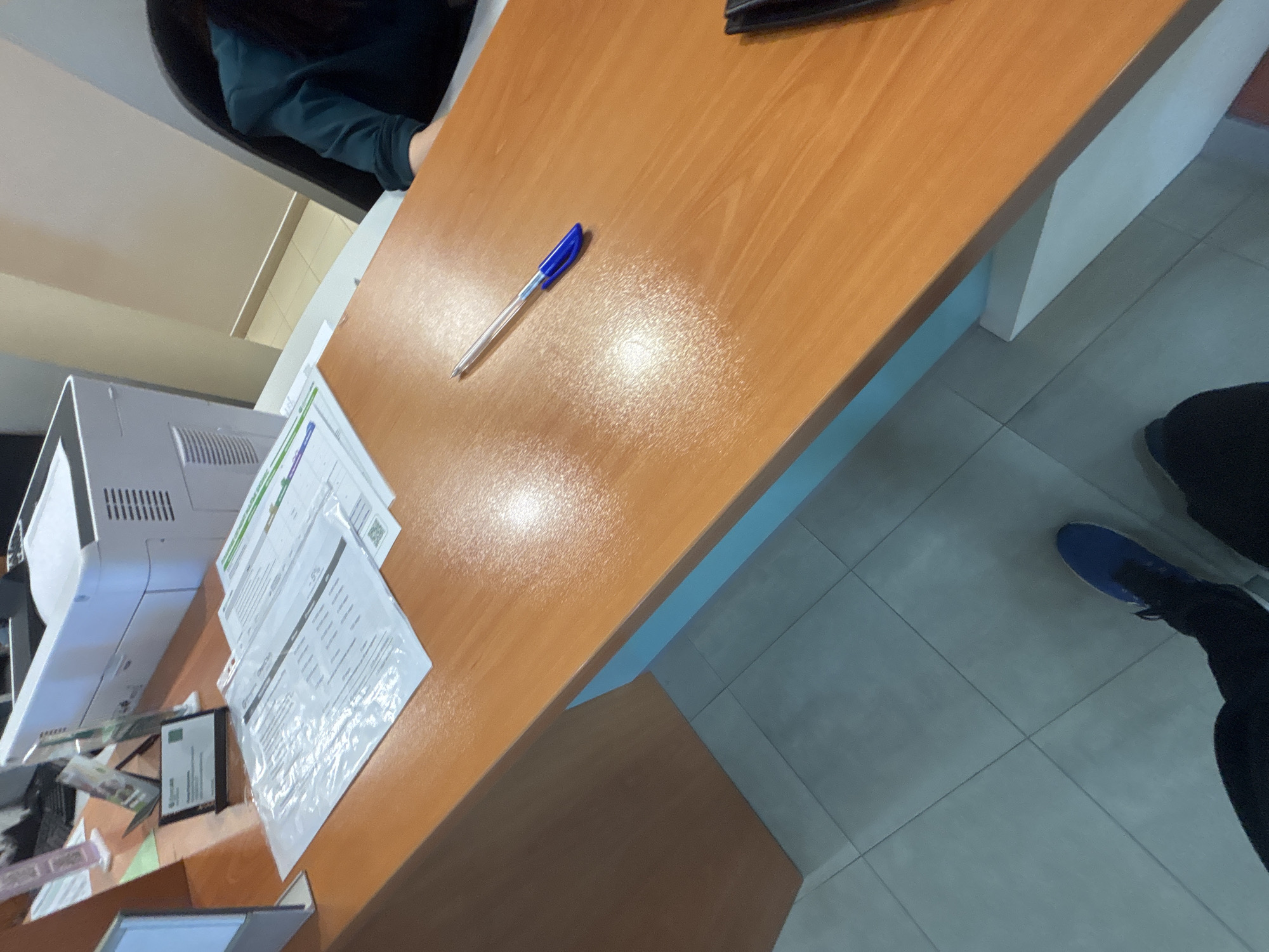The image depicts an office setting in Zlatograd, Bulgaria. A desk made of light brown wood occupies the scene's center. On the desk are several sheets of paper with printed text and a blue ballpoint pen lying diagonally. A printer is partially visible on the left side of the desk. The floor consists of light grey tiles. A person wearing black trousers and blue sneakers is standing on the right side of the desk. Another person is seated to the upper left, wearing a dark blue top. The lighting appears to be artificial, illuminating the desk and surrounding area.