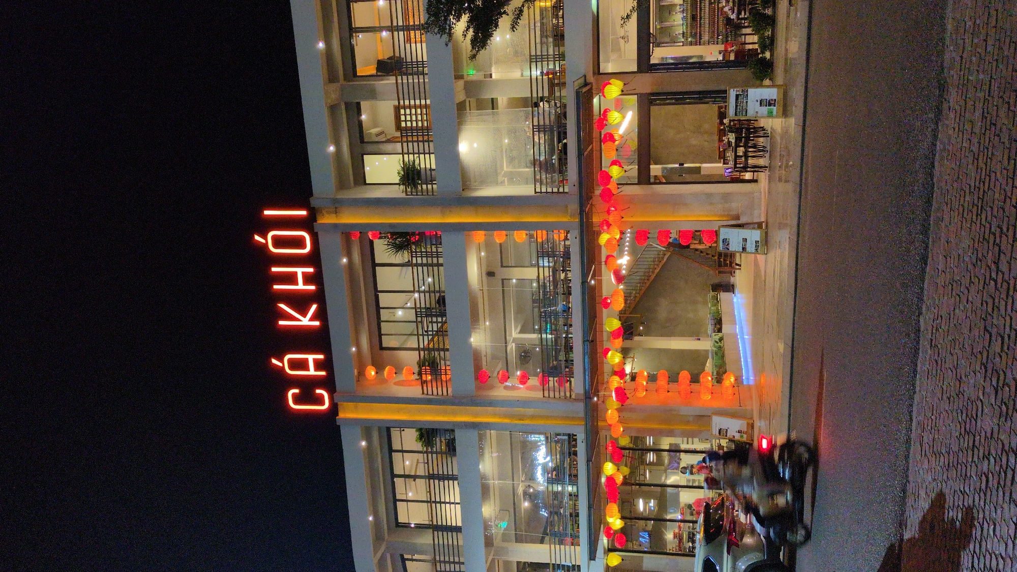 At night, a three-story building stands with bright interior lighting, decorated with hanging lanterns. A vertical neon sign reads "CA KHOI" in red letters. The building's facade is modern, featuring large windows and white pillars. A street runs alongside the building, with a motorcycle and a car visible on the pavement. The location is Quy Nhon Ward, Vietnam, indicated by the Vietnamese sign.