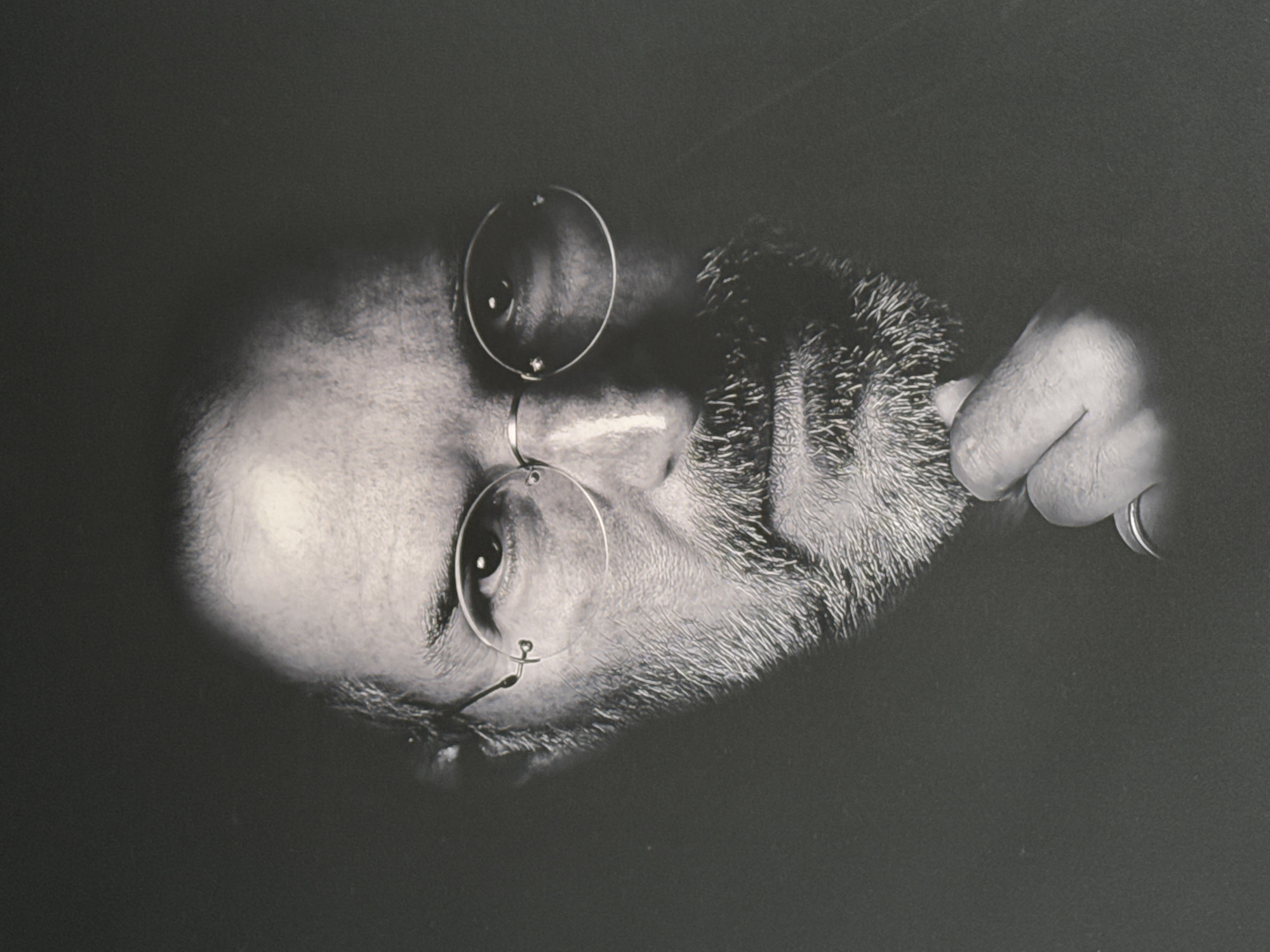 A black and white portrait of a man with a beard and round spectacles is displayed. The man's bald head reflects light on the left side of the image. He is wearing pince-nez style glasses. His hand is partially visible on the right side of the frame, pulling at his beard. The background is dark and indistinct. The image is not taken in the city of Votkinsk, Russia, as there is no indication of that location in the image.