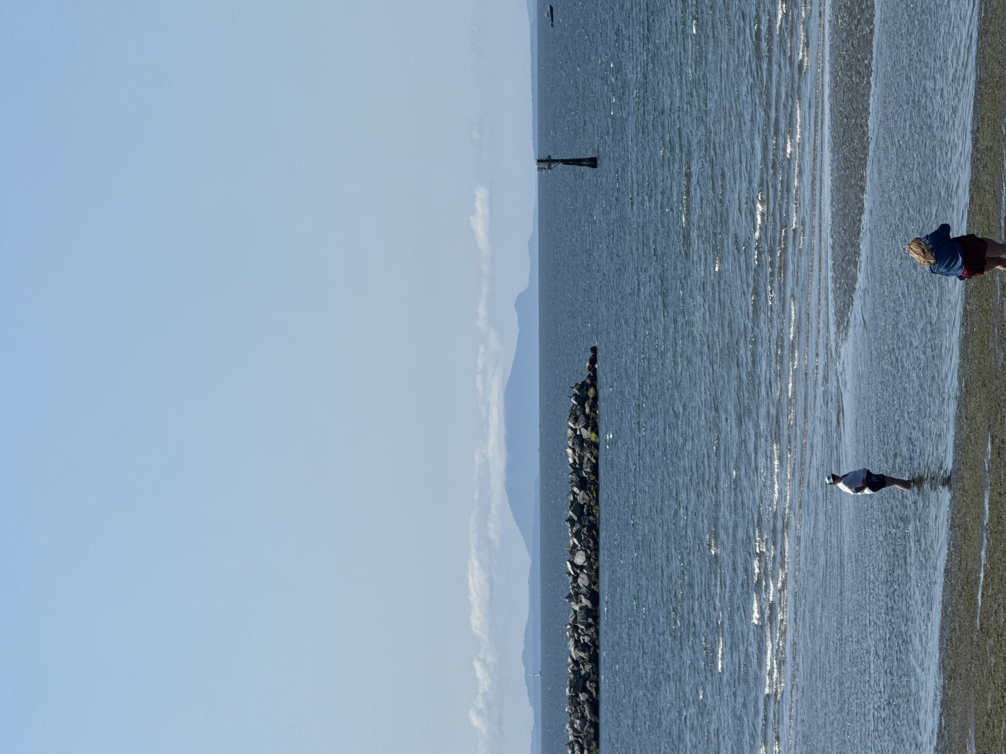 The image depicts a clear, sunny day with a wide expanse of blue sky dominating the upper portion. Below the sky, a calm, blue ocean stretches across the frame. In the distance, a chain of mountainous islands, partially obscured by a soft, white cloud bank, forms the horizon. In the middle ground, a long, dark breakwater constructed of large rocks extends horizontally across the water, creating a barrier. A black, pole-like marker with a rectangular top stands in the water to the right of the breakwater.
Closer to the foreground, the shallow water laps at a sandy and rocky shoreline. Two individuals are present in the water. On the left, a person stands partially submerged in the water, facing away from the viewer, wearing a white t-shirt and shorts and a baseball cap. On the right, another person with blonde hair, wearing a blue jacket and red shorts, is also partially in the water, facing away from the viewer. The water exhibits gentle ripples and the sunlight glints off its surface. The beach area in the immediate foreground is a mixture of sand and dark, wet debris. This scene is consistent with the waterfront area of White Rock, Canada, known for its beaches, breakwater, and views of the southern Gulf Islands.
