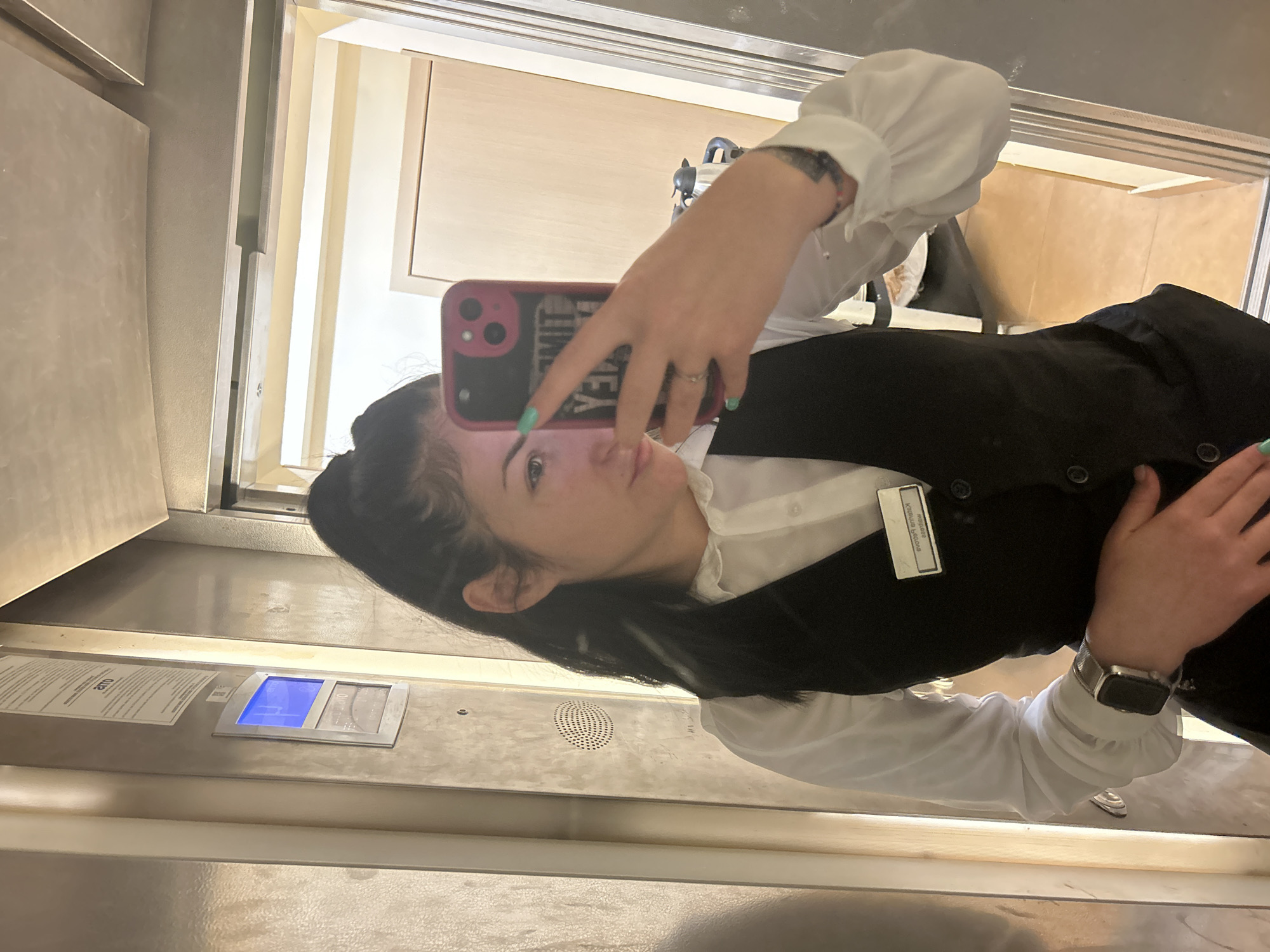 A woman is taking a selfie in what appears to be an elevator. She is wearing a white long-sleeved shirt, a black vest, and a name tag on her vest that reads "Maikela Kostadinova". She is holding a pink phone in her left hand. The elevator has metal panels and a control panel visible.
This location information could not be accurately assessed from the image.