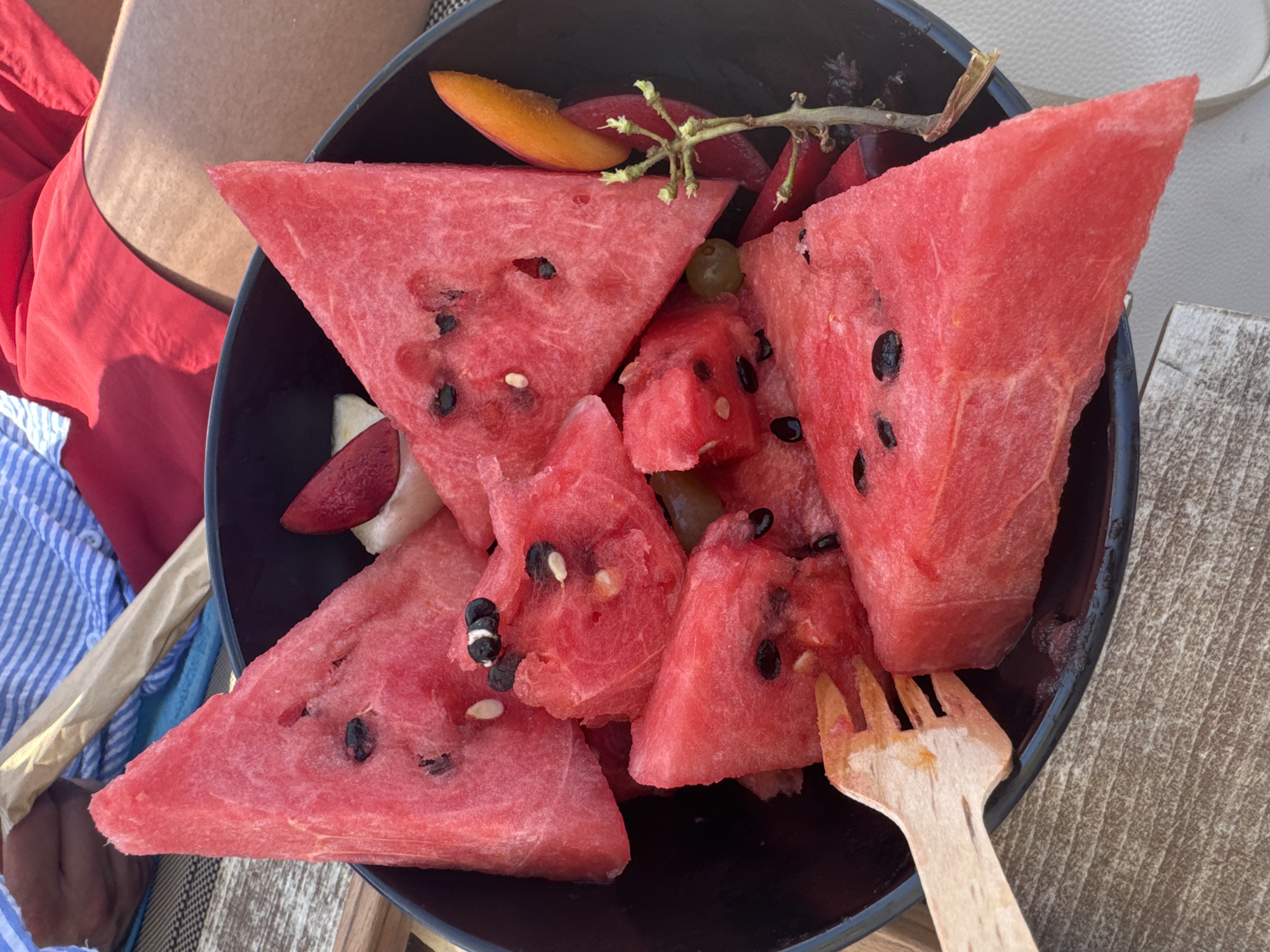 A close-up shot shows a dark bowl overflowing with sliced watermelon. The watermelon pieces are triangular and some are square, with visible black seeds. A light-colored wooden fork is partially submerged in the fruit on the left side of the bowl. In the background, a person's legs are visible wearing red shorts, and a blue and white striped towel or blanket is draped over their lap. The setting appears to be outdoors, possibly near a beach or pool, given the light and the casual attire. The overall scene suggests a refreshing snack on a warm day. The location is in the city of Pallini Municipal Unit, Greece.
