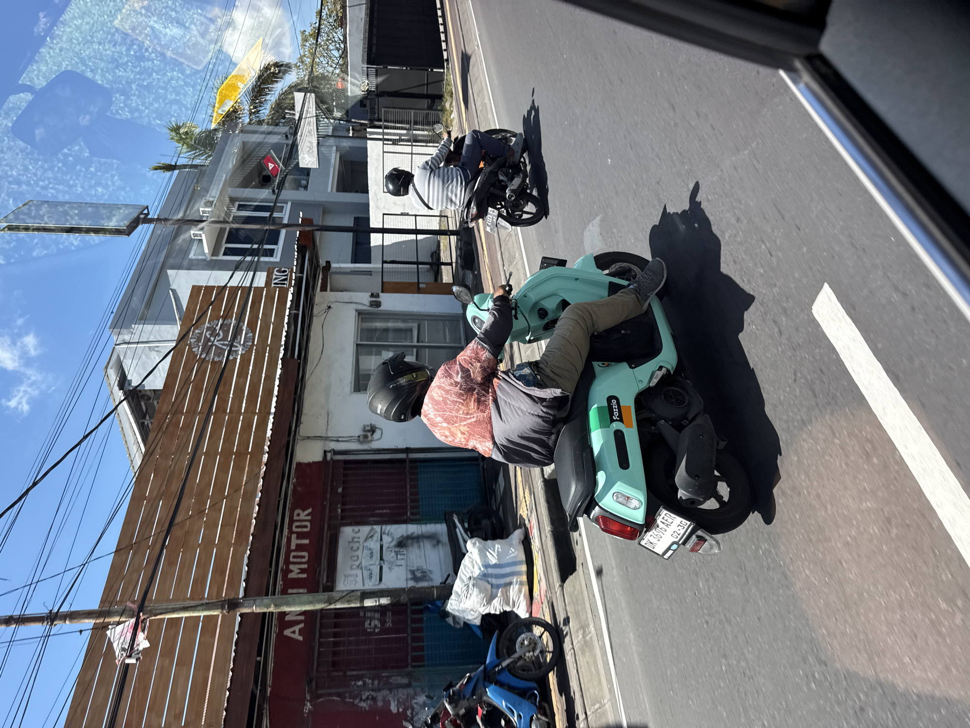 The image captures a vibrant street scene under bright daylight, seemingly taken from inside a moving vehicle due to reflections visible in the upper frame. Two individuals are riding motorbikes on an asphalt road marked with a white line.
In the foreground, a person on a distinctive teal (light blue) Yamaha Fazzio scooter is prominent. The rider wears a black helmet, a patterned reddish-brown long-sleeved shirt, and light-colored pants. The scooter's rear section, including its exhaust and license plate, is clearly visible. The license plate reads "DK 3696 AER" with "02-26" below it, indicating a registration from Bali, Indonesia. Further up the road, another individual rides a black motorbike, also wearing a helmet and a striped shirt.
Buildings line the left side of the street, appearing to be a mix of shops and residences. A prominent sign on one building clearly displays the text "ANI MOTOR". Another partially visible sign includes the letters "ING". Numerous power lines crisscross the sky above the buildings. A blue motorcycle is partially visible, parked near a building on the left. Strong shadows on the road signify bright sunshine, suggesting a midday or early afternoon setting. The visible Indonesian license plate ("DK" for Bali) places the location in Kuta Selatan, Indonesia, consistent with the architecture and street activity.