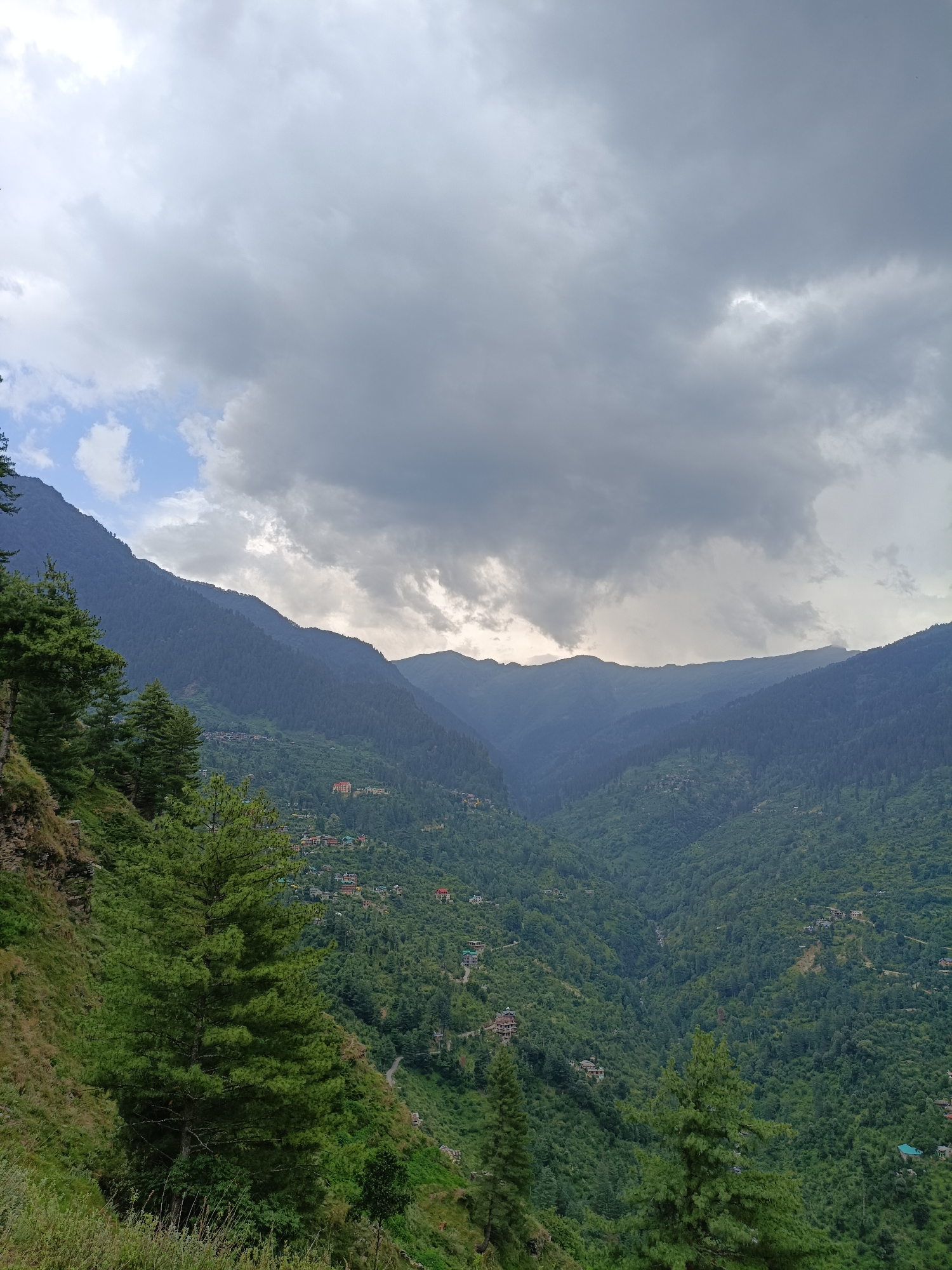 A dramatic, overcast sky looms over a lush, green valley dotted with a scattering of houses and buildings, suggestive of a small village nestled within the mountains. The valley floor is densely covered with trees, creating a textured tapestry of greens that extend up the steep slopes of the surrounding mountains. A winding road is faintly visible, snaking its way through the landscape. In the foreground, the steep, grassy slope of a mountain rises, with several tall pine trees standing sentinel. The clouds are thick and dark, with some lighter patches breaking through, hinting at a recent or approaching rain. The overall mood is one of natural grandeur and a sense of peaceful isolation. The scene appears to be captured in the daytime, given the light. The location is identified as Chhaki, India.