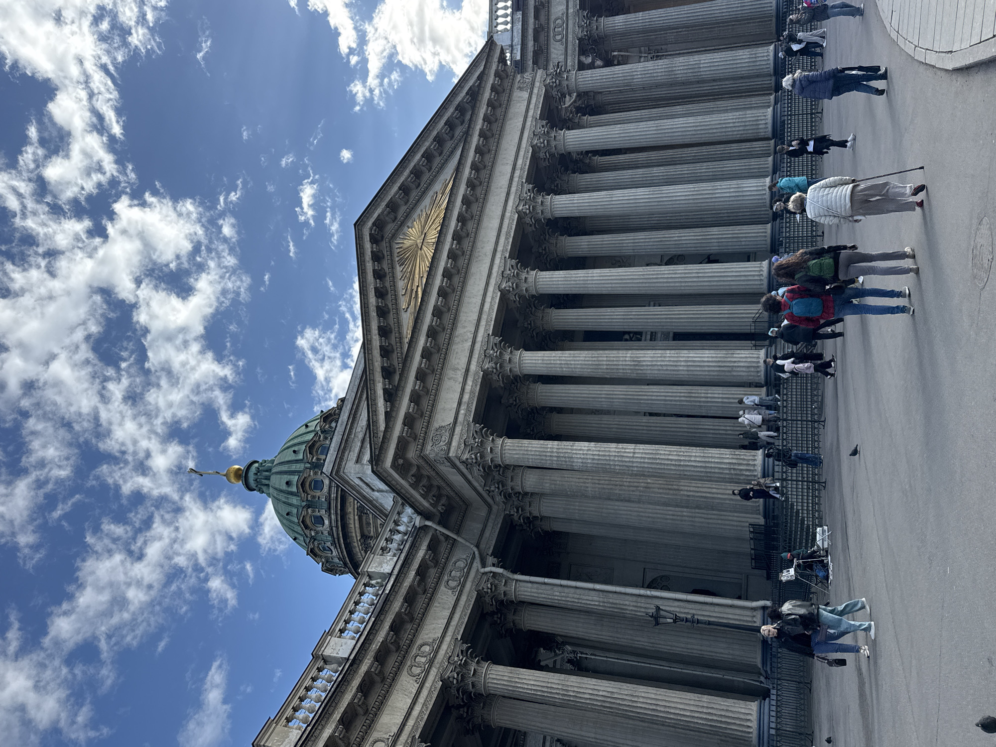 The image shows the Kazan Cathedral in Saint Petersburg, Russia, seen from a low angle. The cathedral's dome and a series of columns dominate the frame. Several people are walking along the street in front of the cathedral. The sky is blue with scattered clouds.