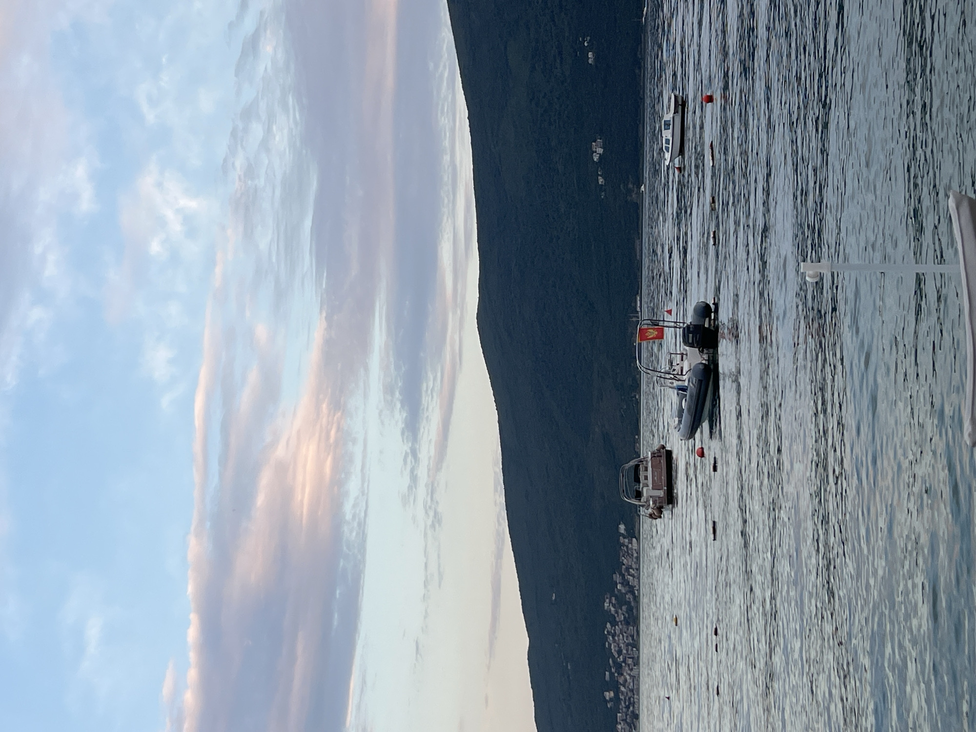 The image depicts a body of water with several boats, a dark, forested mountainside, and a hazy sky with wispy clouds. The orientation of the image is vertical, as if it were taken on a phone and rotated. The sky transitions from a pale blue to a lighter, almost white hue, with streaks of pinkish-orange, suggesting either sunrise or sunset. The water is choppy with a strong texture of ripples and reflects the light in shimmering patterns.
Several boats are visible on the water. Two prominent ones are closer to the foreground. One appears to be a rigid inflatable boat (RIB) with an outboard motor. It has a flag with red and yellow colors. Another boat, possibly a smaller vessel or dinghy, is nearby. Further out in the water, a white boat with windows is anchored. There are also a few buoys visible in the water, one of which is red.
The dark, steep mountainside forms a stark contrast with the sky and water, and it appears to be covered in dense vegetation. A scattering of buildings or houses can be seen nestled on the lower slopes of the mountain, suggesting a coastal settlement.
The overall impression is a serene coastal scene, likely captured during the golden hour due to the soft, warm light in the sky. The location appears to be a bay or harbor, with the mountains providing a dramatic backdrop to the tranquil waters. No people are explicitly visible in the image. The time of day appears to be either early morning or late evening, characterized by the soft lighting and the colors in the sky. The weather appears to be clear and calm, despite the ripples on the water.