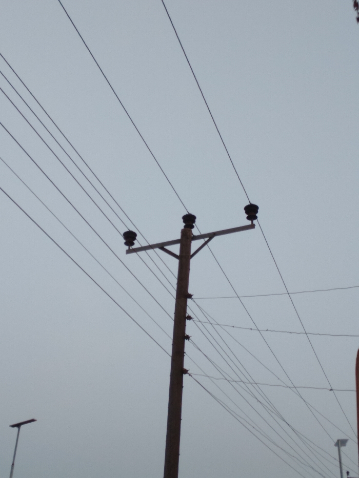 The image presents a view of a utility pole and an intricate network of overhead power lines set against a uniformly overcast, light gray sky, consistent with an outdoor scene in Ejule, Nigeria.
A prominent utility pole, likely made of wood or concrete, stands vertically in the mid-right portion of the frame. Near its top, a horizontal cross-arm extends, supporting three distinct black, spool-like insulators. From these insulators, three main power lines extend across the upper part of the frame, stretching diagonally from left to right.
Numerous other, thinner electrical wires crisscross the sky, both connected to the main pole at various points (including smaller insulators lower down) and extending out of the frame in multiple directions, creating a dense web. This suggests a complex electrical distribution system.
In the bottom-left corner, the top of a modern streetlight or similar tall pole is partially visible. A shorter pole, possibly another streetlight, is also faintly discernible in the bottom-right background. The consistent grayness of the sky indicates an overcast day, suggesting diffused light conditions without direct sunlight. There are no people visible, and no specific activities or events are taking place. No text is visible anywhere in the image.