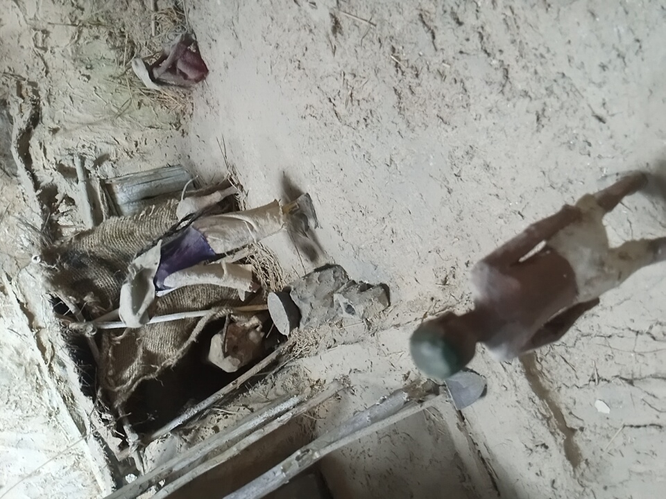 The media file shows a scene with two apparent human figures, likely mannequins or dolls, in a dimly lit, rustic setting. One figure is lying down, partially covered by a burlap-like material and what appears to be fabric clothing in shades of purple and white. It is positioned in what looks like a shallow pit or crevice within the earthy surroundings. The other figure is standing, facing away from the camera, and is also composed of a dark material with lighter patches, possibly representing skin or clothing. Its head is topped with a dark green object.
The ground is uneven and appears to be made of dirt and straw, with some debris scattered around. There are hints of rough wooden structures and possibly some discarded objects like a piece of tire in the background. The overall impression is of an abandoned or neglected space.
The lighting is subdued, suggesting it might be indoors or outdoors at dusk or dawn, or in an area with limited light sources. There are no clear indicators of specific weather conditions. The environment suggests a rural or perhaps an undeveloped area, consistent with the provided location context of Angren, Uzbekistan, which is known for its industrial and mining activities, but also has rural surroundings.
The figures' poses and the general state of the scene do not convey any specific activity or event. They appear to be static. There is no visible text in the image. The emotion evoked by the scene is one of stillness, abandonment, and perhaps a touch of desolation due to the rough materials and dim lighting.
