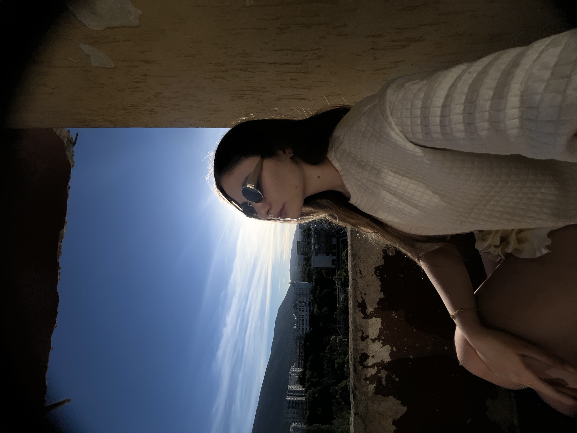 A young woman with long, dark hair and sunglasses is sitting on a balcony, looking towards the sky. She is wearing a textured white sweater. The background shows a cityscape with buildings and mountains under a clear blue sky with some wispy clouds. The sun is visible, creating a lens flare and casting a warm glow on the scene. The balcony railing and wall show signs of wear and tear. The image appears to be taken in the late afternoon or early evening, judging by the lighting.