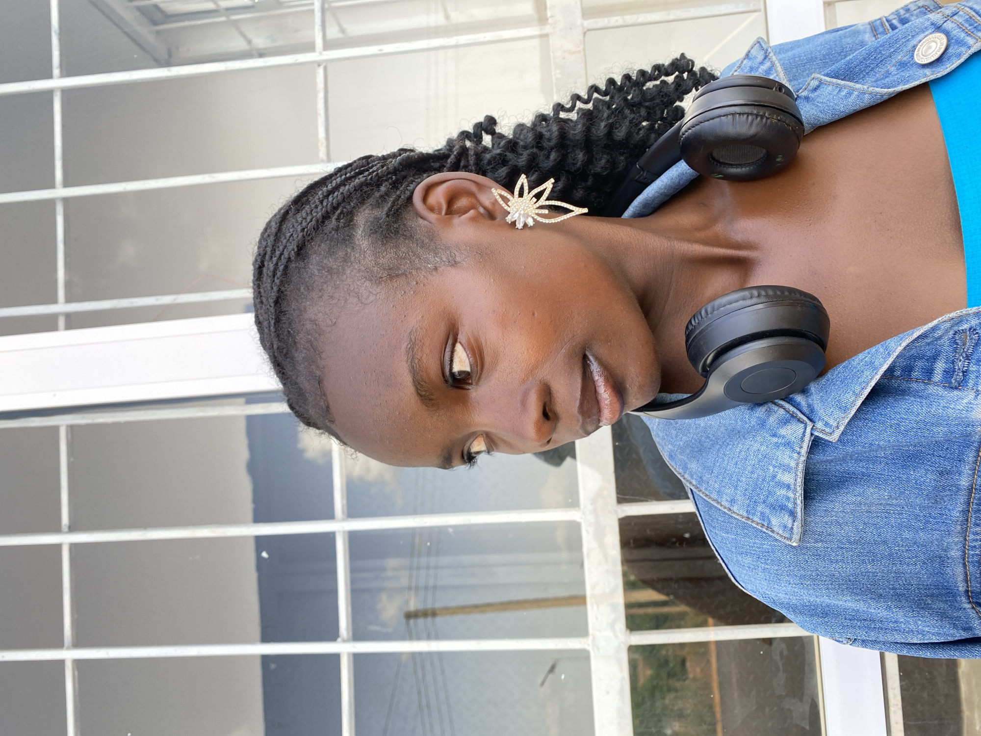A medium close-up shot shows a Black female subject from the chest up, looking to her left. She has braided hair, a prominent gold and diamond earring in her left ear, and black headphones draped around her neck. She is wearing a blue denim jacket over a bright blue top. The background features a white-framed window with multiple panes, reflecting the sky and some vegetation. The image was taken in Bar Jirgi Summa, Nigeria.