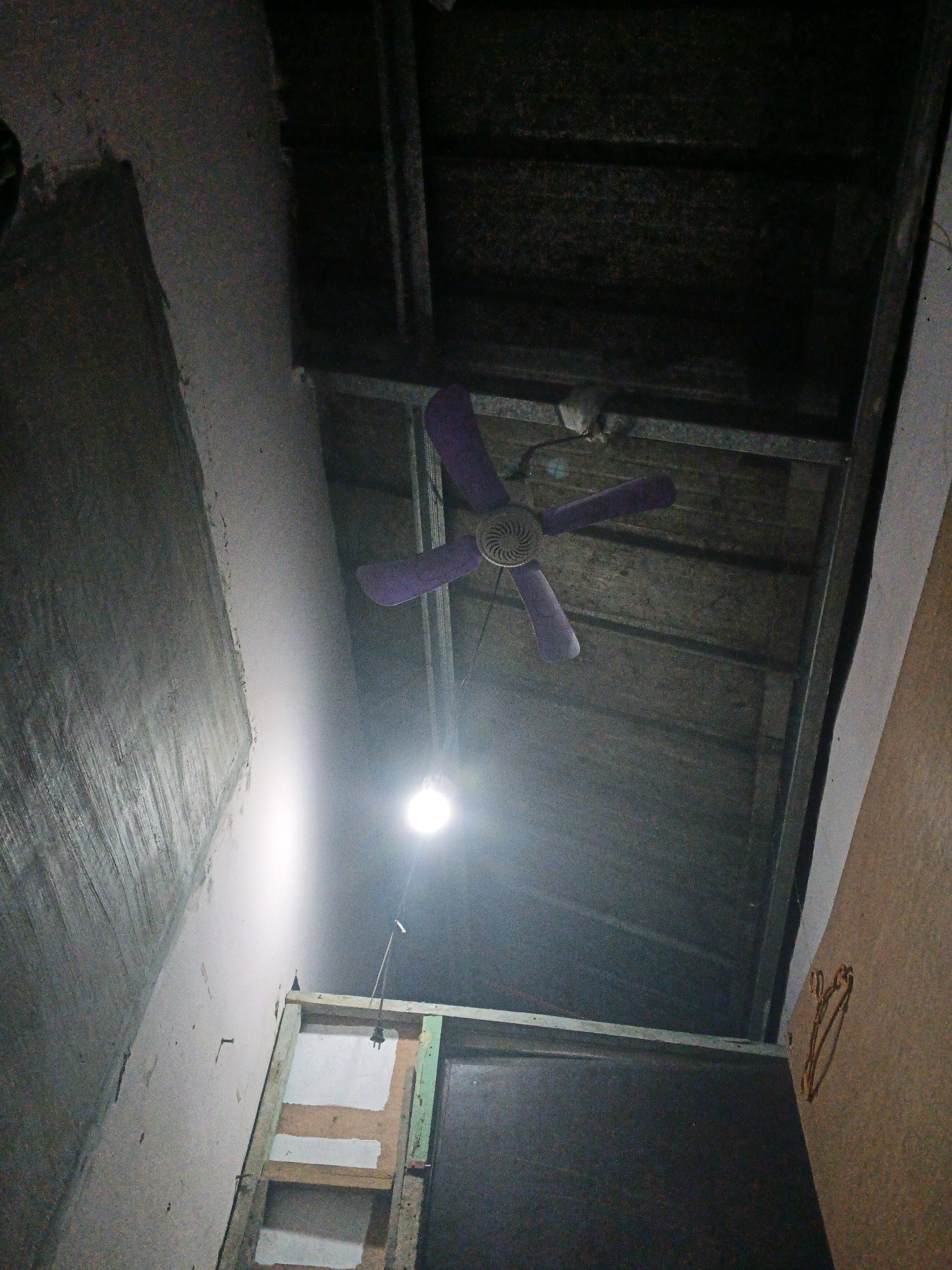 An overhead shot shows a purple ceiling fan with three blades attached to a metal frame that supports a dark, textured ceiling. Below the fan, a bare light bulb hangs from a wire, emitting a bright, direct light. To the left, a roughly finished wall with a textured, gray surface extends upwards. Below this wall, a section of exposed wooden framing is visible, with what appear to be white panels inserted within the structure. To the right, a plain, tan-colored wall is present. The image captures an indoor, unfinished or under-construction environment. This scene appears to be located in Santa Barbara, Philippines, a municipality within the province of Iloilo, Western Visayas region.