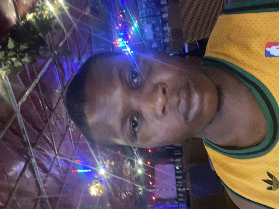 A man is facing the camera, illuminated by multicolored lights. He wears a yellow and green basketball jersey with an NBA logo. Behind him is a structure, possibly a decorative cage, adorned with more lights and greenery. His skin has a sheen, and his expression is neutral as he looks directly into the lens. The composition suggests an outdoor setting or a space decorated for an event or festivity.