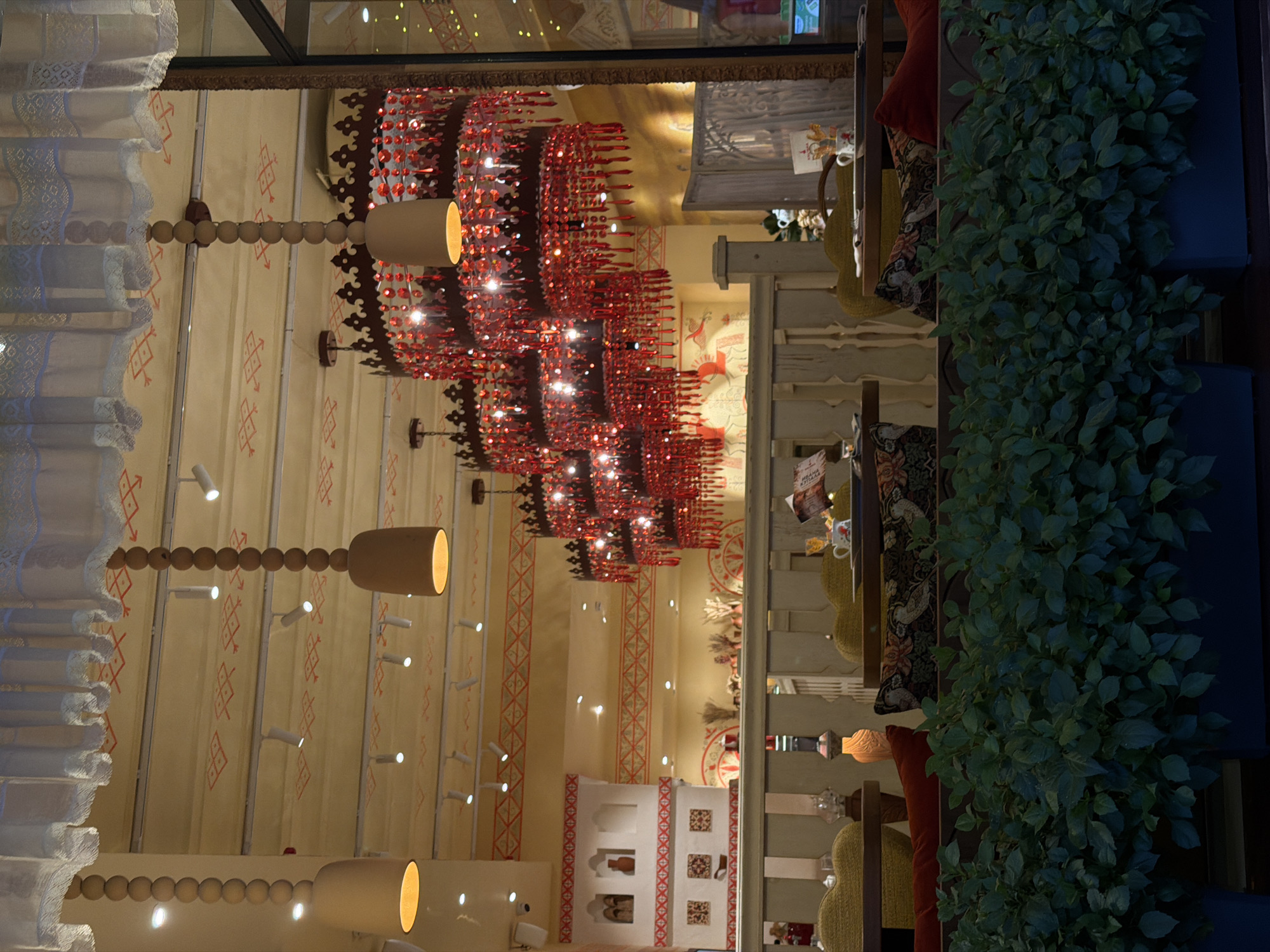 The image shows the interior of a restaurant, likely in Moscow, Russia, with a dimly lit and warm ambiance. The scene is dominated by decorative elements and architectural features. A large, ornate chandelier made of red, crystalline-like pieces hangs prominently in the center, casting a warm glow. To the left, white lace curtains with intricate patterns frame the scene, adding a touch of elegance. The walls are decorated with a cream-colored fabric adorned with a repeating pattern of red geometric symbols.
Several pendant lamps, with cylindrical shades and exposed bulbs, are visible, contributing to the warm lighting. Along the ceiling, small spotlights are strategically placed. A tall, dark green plant cascades down a dark blue planter on the right side of the image, adding a touch of nature to the indoor setting.
In the background, glimpses of furniture, including upholstered chairs and cushions with patterned covers, suggest a comfortable dining area. A small, white architectural structure resembling a miniature building with decorative squares is also present, adding a unique visual element.
The lighting suggests it is either evening or night, and the overall mood is cozy and inviting. There are no visible people in the image, focusing the attention on the setting and its decor. The image is oriented vertically, giving a sense of height and depth to the restaurant's interior. No discernible text is clearly visible.