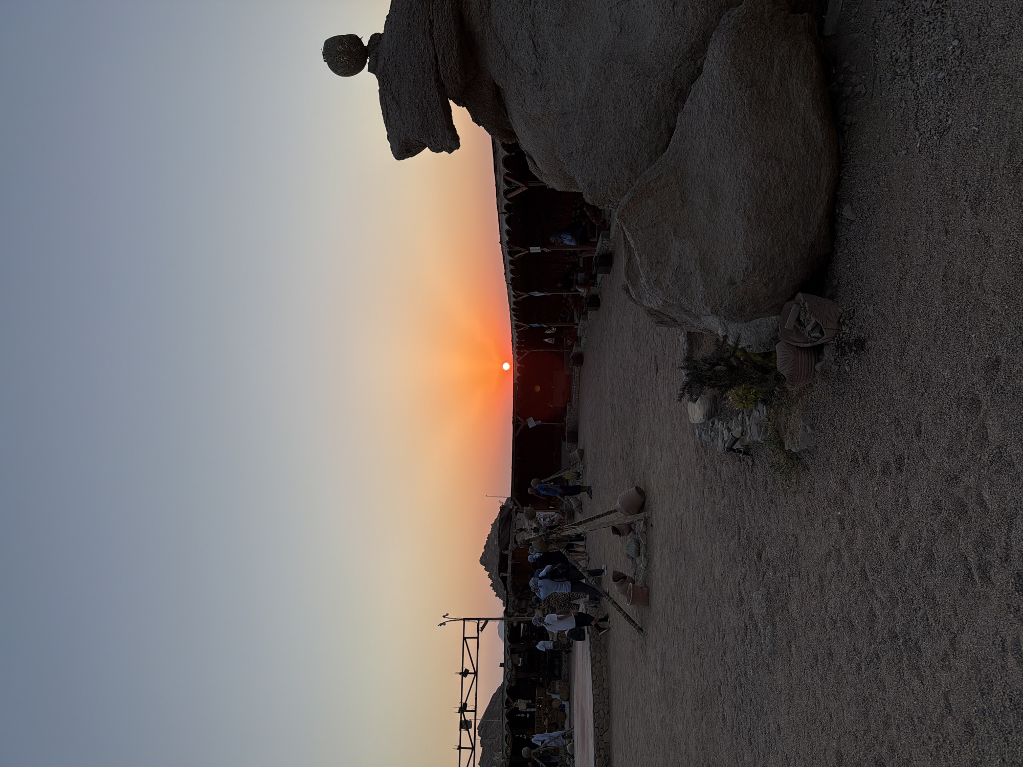 The image depicts a desert setting in Sharm El Sheikh, Egypt, during sunset. The sky transitions from a light blue at the top to shades of orange and pink near the horizon, where the sun is setting. In the foreground, there is a sandy ground scattered with small pebbles and rocks. To the right, a large, irregularly shaped rock formation dominates the scene, with a smaller, rounded rock balanced on top of it.
A wooden structure with a thatched roof runs horizontally across the middle ground, serving as a shaded seating area. Several people, appearing to be tourists, are visible in and around this area, some standing and others seated. They are dressed in casual clothing, and some are wearing headscarves.
In the left and middle ground, there are more structures, possibly market stalls or food stands, with tents and wooden elements. Various objects are scattered around the area, including earthenware pots, wooden poles forming a tripod, and what appears to be a small fire pit. In the far left, a stage or platform is visible. The overall impression is of an outdoor entertainment or cultural venue set against a desert landscape.