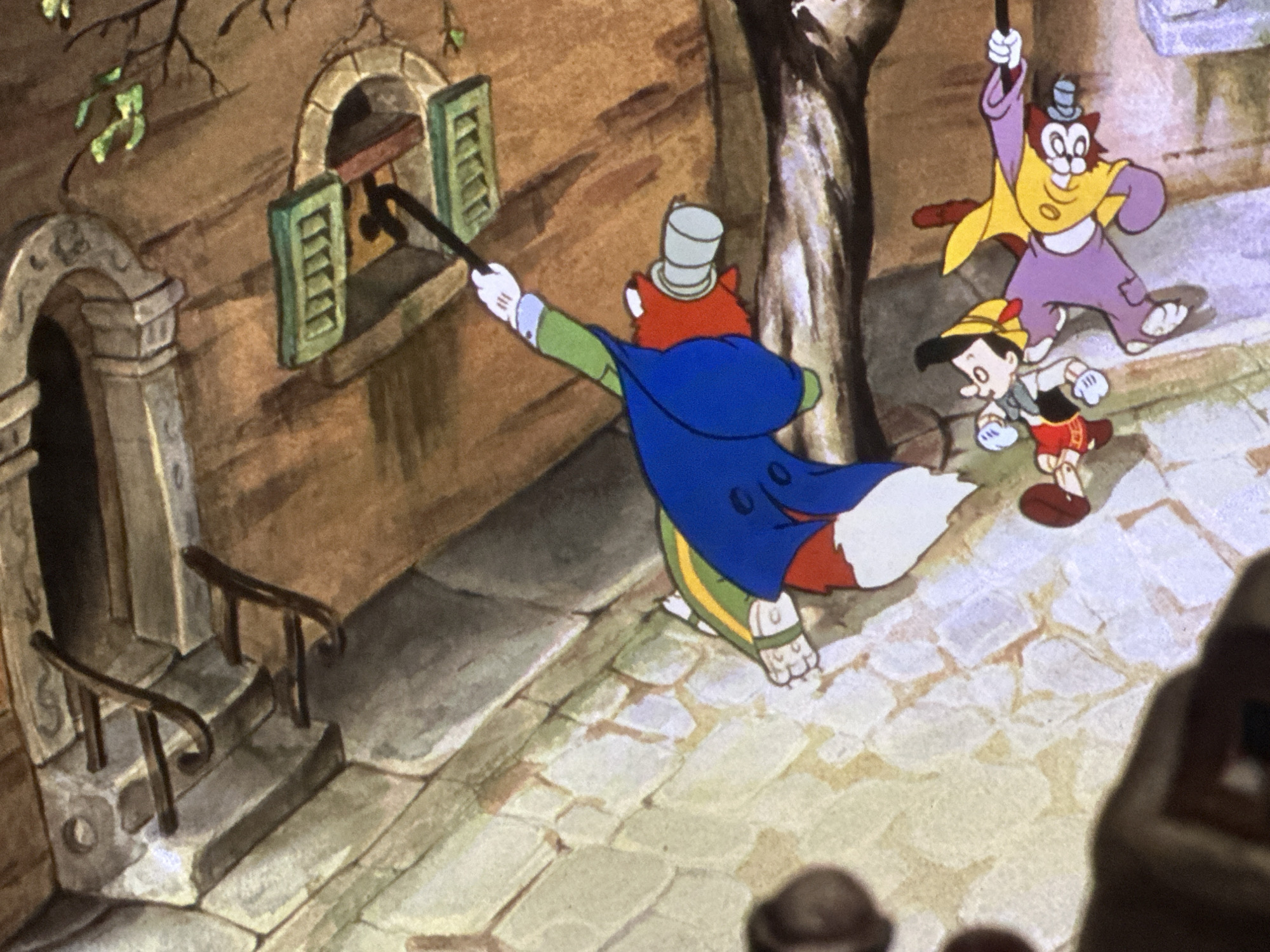The image captures an animated street scene from a classic cartoon, featuring three main characters on a cobbled pathway beside an old building. In the center-left, a fox-like character, identifiable as Honest John, wears a blue cape, green attire, white gloves, and a grey top hat. He is actively using a long black cane to poke at the closed green shutters of a window on the stone building.
Behind Honest John, a cat-like character, likely Gideon, is shown clinging to the upper part of the same cane. Gideon is dressed in a purple jumpsuit, a yellow cape, and a small light blue top hat. To the right of Gideon, Pinocchio, the wooden puppet, is visible, wearing a yellow hat with a feather, a white shirt, black vest, and red shorts.
The setting is an old European town, characterized by sturdy stone architecture. An arched doorway on the left leads to a building, preceded by a short set of stone steps with simple metal railings. The street surface is composed of uneven cobblestones. A dark, textured tree trunk rises on the right side of the frame. The scene is brightly lit, indicating it is daytime. The activity suggests an attempt by Honest John and Gideon to gain access or attention from within the building, with Pinocchio present alongside them. There is no visible text in the image.
