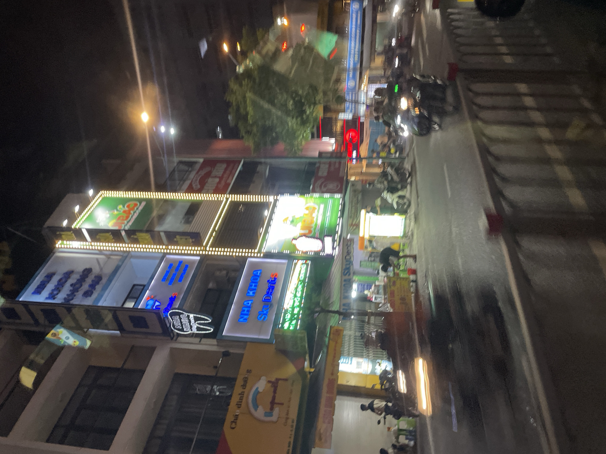This is a night-time, vertical-oriented photograph of a street scene in Thủ Đức, Vietnam. The image is somewhat blurry and appears to be taken from a moving vehicle. The scene depicts a street lined with multi-story buildings, many of which have illuminated signs.
The subjects include several motorbikes parked along the street and in front of the buildings. Some people are visible on or near the motorbikes. The buildings house various businesses, indicated by the illuminated signs. One sign reads "NHA KHOA SKY DENT'S" in blue letters with a white outline, suggesting a dental clinic. Another sign has green lettering and a circular logo. A yellow sign with red and blue text is also visible.
The time of day is clearly night, with artificial lights from signs and street lamps illuminating the scene. The weather is not explicitly discernible but the lack of rain suggests it might be clear. The presence of motorbikes and the style of the buildings and signage are strong indicators of a location in Vietnam. The overall atmosphere is that of a busy commercial street at night. The blurriness of the image might convey a sense of movement and the dynamic nature of urban life.
