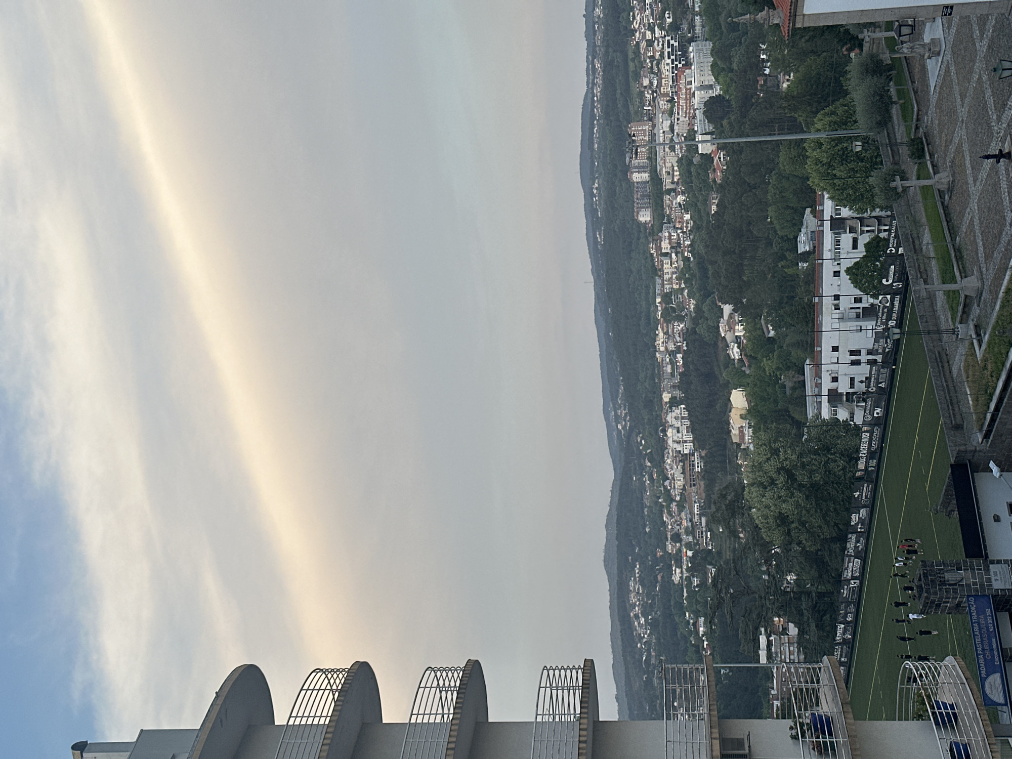 The image depicts a cityscape of Vila Real, Portugal, under a sky with diffuse sunlight. The foreground shows a partial view of a building with architectural elements including concrete arcs and metal fencing. In the middle ground, a green sports field with players practicing is visible. Advertisements border the field. Beyond the field lies a densely populated hillside with buildings interspersed among trees. In the upper portion of the image, the sky presents a hazy appearance with subtle color variations. The city sprawls over the landscape. A sign advertises: Padaria Pastelaria Tradição Churrasqueira.