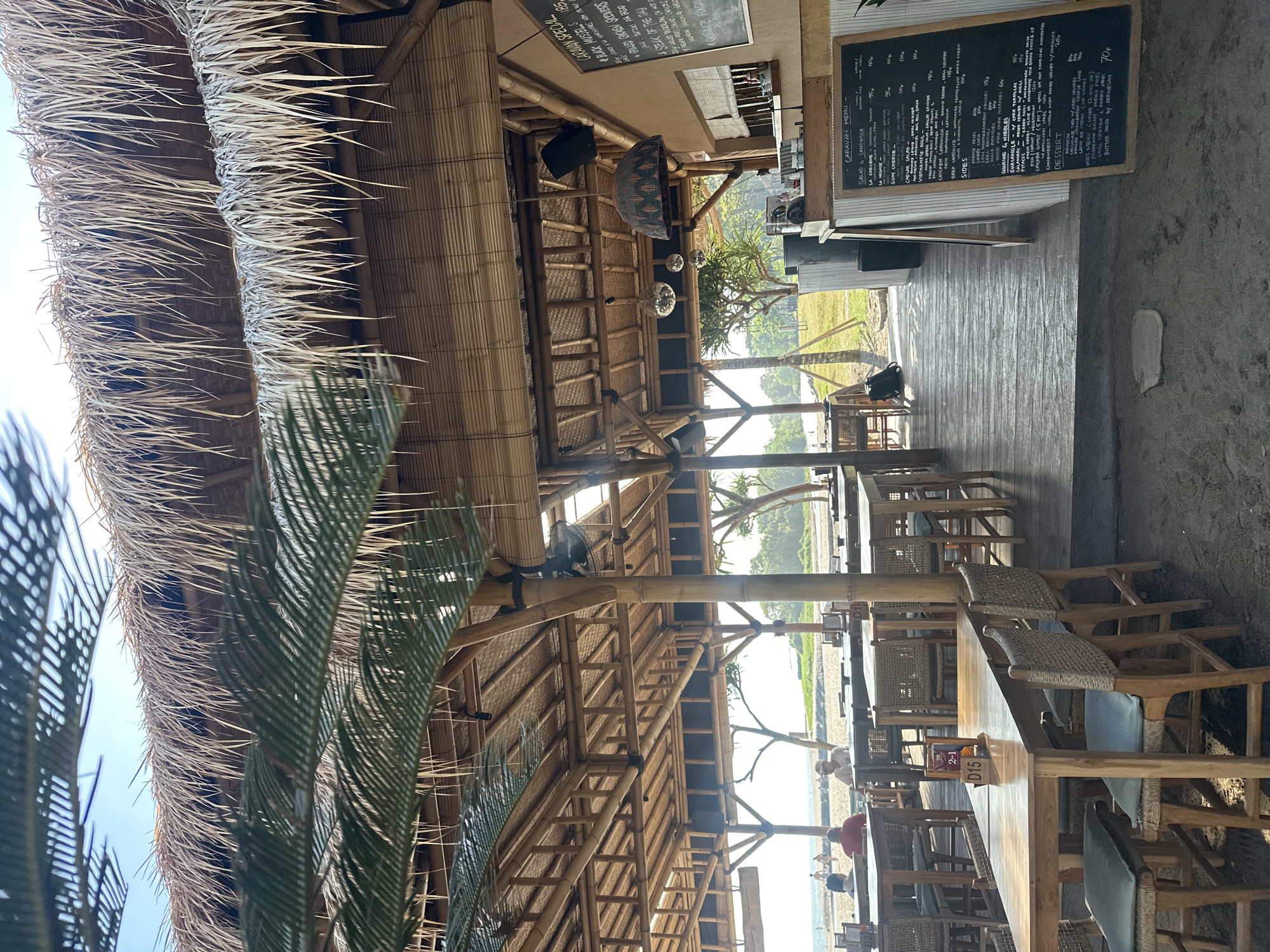 The image depicts an outdoor dining area of a beachfront restaurant in North Kuta, Indonesia, likely at the "Caravan" establishment given the menu board. The structure is built with natural materials, featuring a thatched roof and bamboo supports. Tables and chairs, made of light-colored wood and woven rattan, are arranged on a wooden deck. In the foreground, several tables are visible, some with cushioned seats. In the background, the sandy beach and ocean are present, with a few people casually strolling. A black chalkboard menu is prominently displayed on the right side of the image, listing various food and drink items. The overall atmosphere is relaxed and tropical, suggesting a daytime scene.