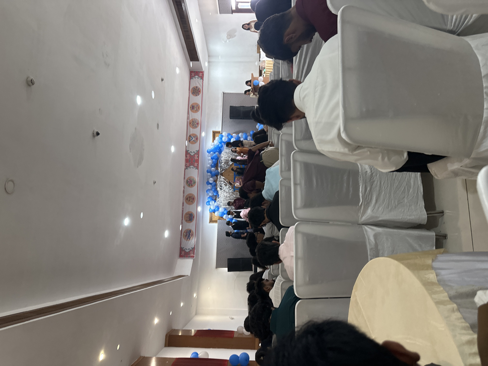 The image depicts an indoor event, likely a ceremony or gathering, set in a hall. The ceiling is high and white, with recessed lighting. A decorative banner featuring circular motifs is displayed along the top of a wall at the far end of the hall. The space is furnished with numerous white chairs, many covered with white fabric. In the foreground, several people are seated, viewed from behind. The lighting is bright and appears to be from overhead fixtures. The overall atmosphere suggests a formal or semi-formal occasion.
