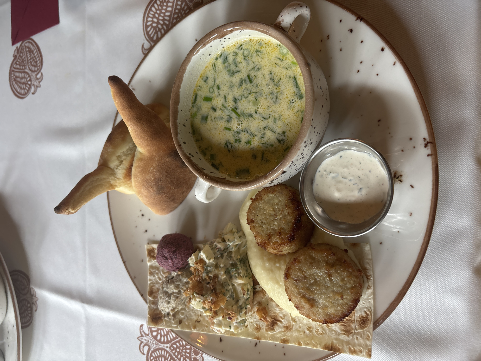 A top-down view of a plate of food is presented on a white tablecloth with a subtle pomegranate motif. The plate contains a creamy soup in a speckled ceramic bowl with a handle, garnished with fresh green herbs. To the left of the soup is a piece of golden-brown bread shaped like a crescent moon or a horn. Below these items, arranged on a flatbread, are two golden-brown, circular patties, likely made from ground meat or vegetables, alongside a dollop of mashed potatoes. Further to the left on the flatbread are a small mound of a dark purple substance, perhaps a beet puree, and a salad composed of chopped vegetables and herbs, topped with crispy fried onions. A small metal ramekin filled with a creamy, herb-flecked sauce sits to the right of the soup. The setting appears to be a restaurant, suggested by the tablecloth and plating, and the meal is likely served during daylight hours. The food appears fresh and appetizing, indicating a pleasant dining experience. There is no visible text in the image.