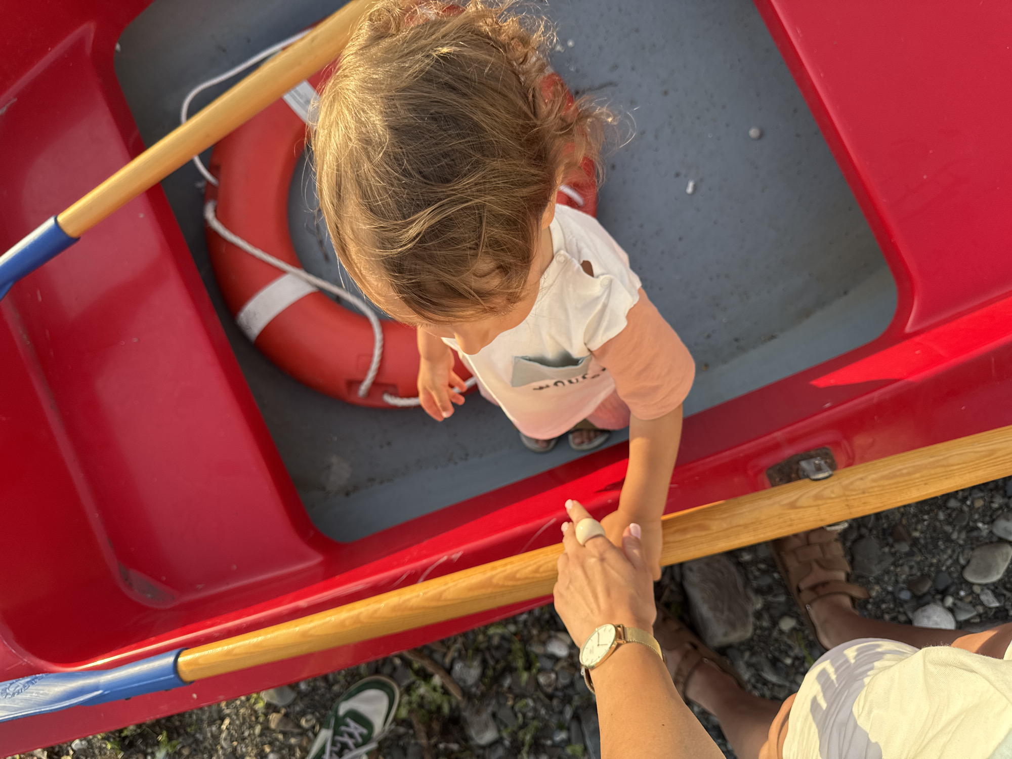 In Sochi, Russia, a child with light brown hair sits in a red boat, wearing a white t-shirt and peach-colored shorts. A person's arms are visible, one wearing a gold-banded watch, holding a wooden oar. A red life preserver with white stripes is also in the boat. The boat is on a shore with pebbles and some green plants.