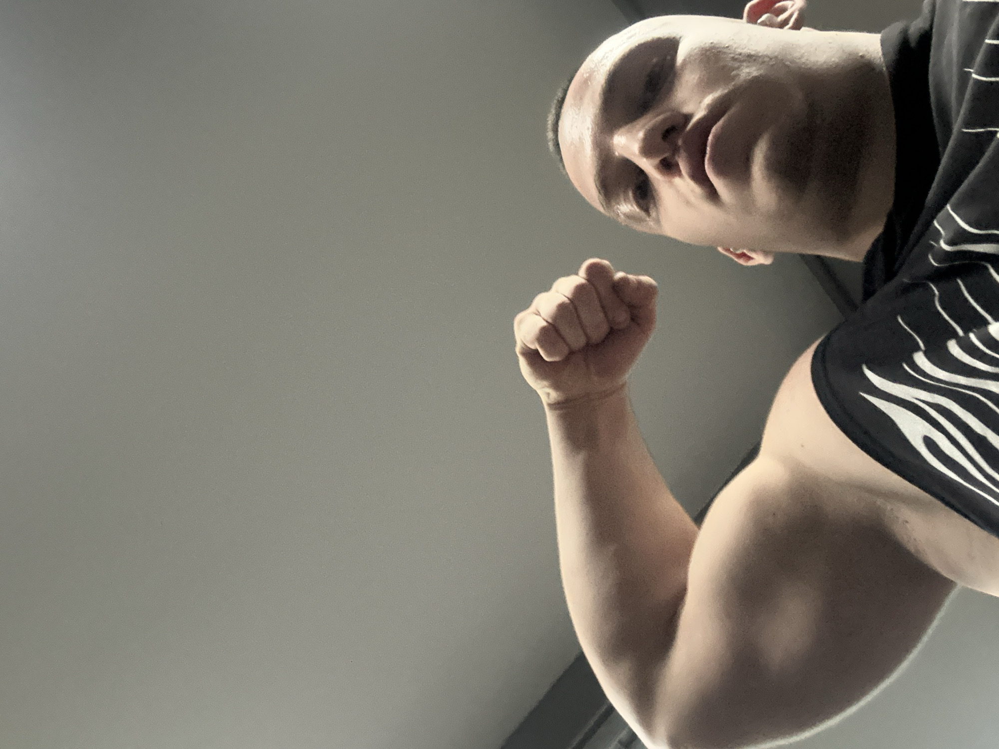 A low-angle shot shows a muscular man with a shaved head flexing his right bicep. He is wearing a black t-shirt with white curved lines. The background is a plain grey ceiling. There is no evidence to suggest the location is Burgas, Bulgaria.