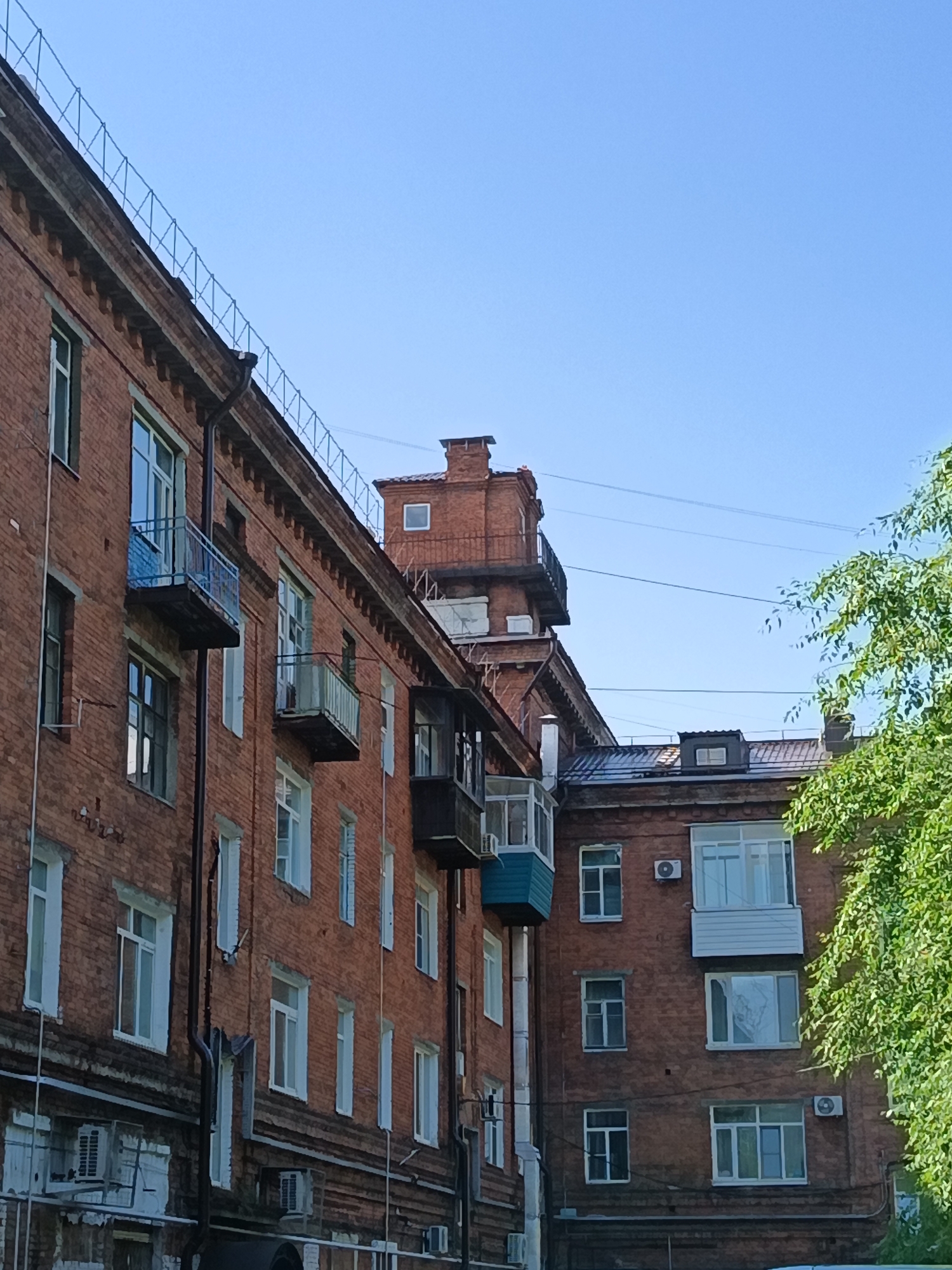 This is a photograph of a brick apartment building in Komsomolsk-on-Amur, Russia. The building has multiple stories with numerous windows and balconies. The architecture appears to be Soviet-era. There is a clear blue sky and some green foliage is visible on the right side of the frame. No people are visible in the image. The image is taken from a slightly low angle looking up at the building. The building's facade is made of reddish-brown bricks. Some of the balconies have metal railings. There are also air conditioning units mounted on the exterior walls. Power lines are visible against the sky.