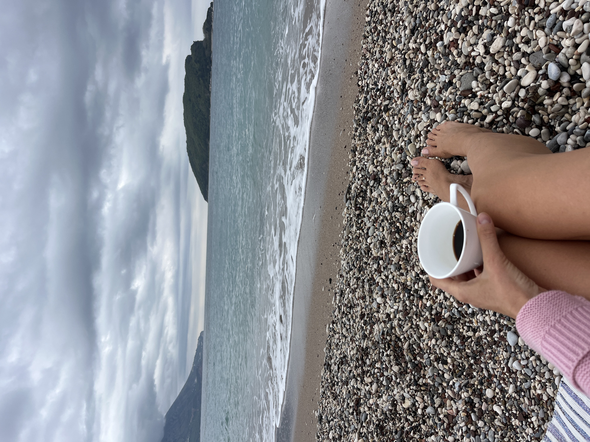 The image, which appears rotated 90 degrees counter-clockwise from its intended horizontal view, captures a serene pebble beach scene in Budva, Montenegro, from a first-person perspective. In the foreground, the bare legs and feet of an individual rest on the smooth, multi-colored pebbles of the beach. The person is holding a plain white mug, containing a dark beverage, in their hands. A sleeve of a light pink knitted sweater and a blue and white striped fabric are also visible, suggesting they are dressed warmly for the weather.
The pebble beach stretches along the coastline, meeting the calm, greyish-blue waters of the sea. Gentle white-capped waves are seen breaking softly at the shoreline. In the mid-ground, a large, dark green, mountainous landmass covered in foliage rises prominently from the sea, receding into the distance. The sky above is completely overcast with thick, grey clouds, indicating a cloudy or moody day. The overall impression is one of peaceful relaxation and quiet contemplation by the sea, despite the overcast conditions. No discernible text is present in the image.