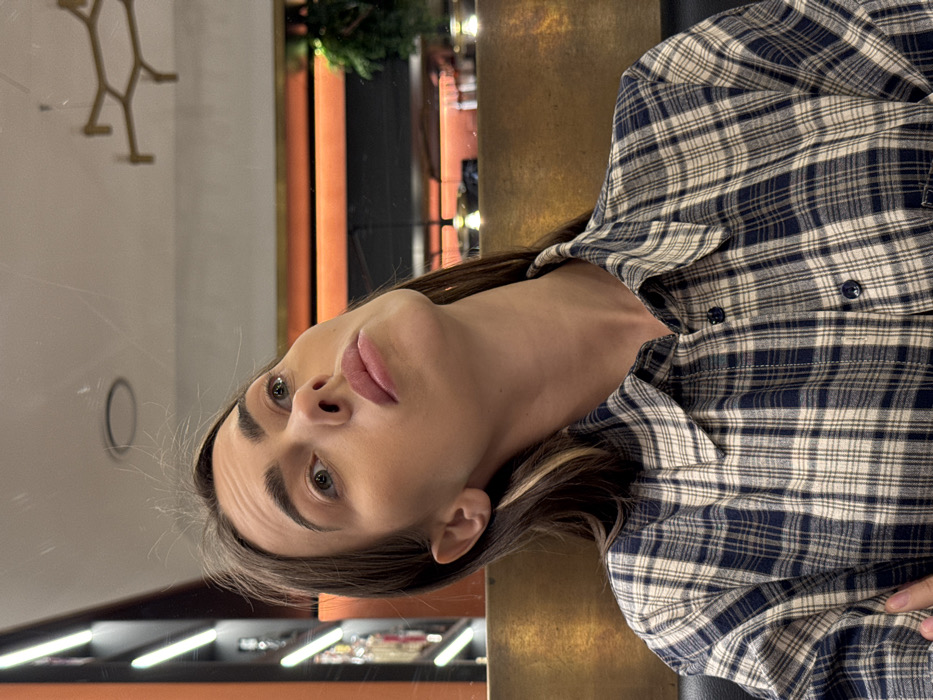 A female individual is depicted in a chest-up shot, seated indoors within an establishment in Verona, Italy. She has dark brown hair with visible lighter highlights and light-colored eyes. She is wearing a blue and off-white plaid button-up shirt. Her head is tilted upwards and to her right, with her gaze directed towards the upper right of the frame, maintaining a neutral expression. The background is out of focus, indicating a modern interior. A vertical structure with strip lighting is visible on the left. A golden-toned horizontal panel separates the subject from the mid-ground, which features a horizontal band of orange lighting. Reflections of light sources are visible on a surface below the orange lighting. The ceiling reflects a circular light fixture and an angular, golden geometric light fixture. Green foliage is partially visible on the far right.