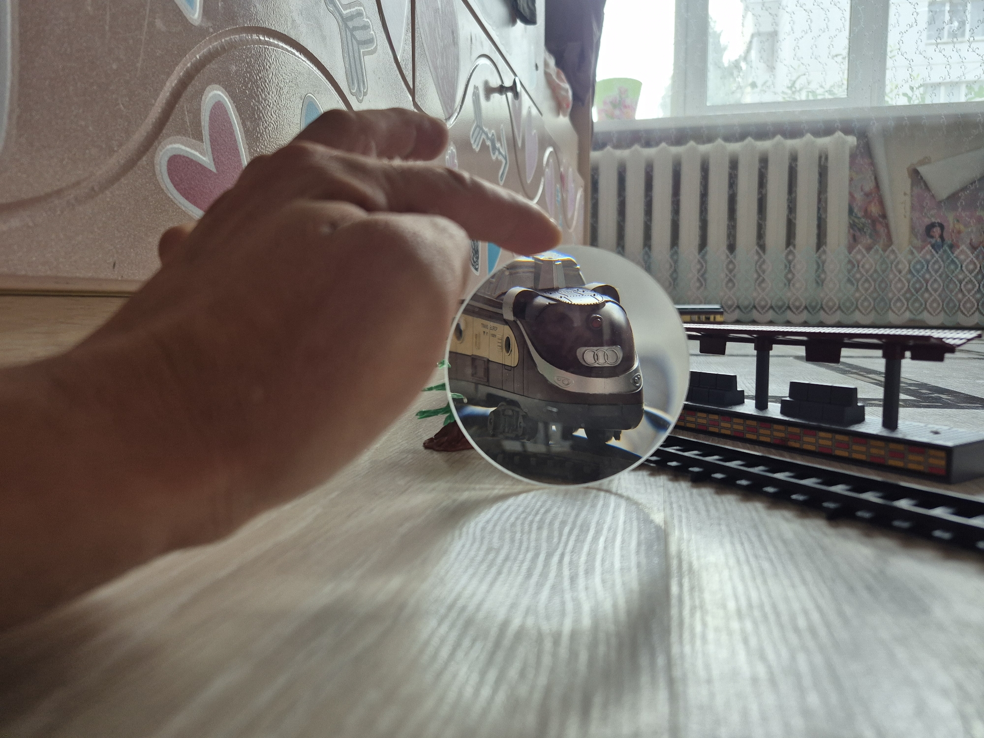 An indoor shot captures a hand holding a magnifying glass over a toy train. The train is dark brown with silver accents and sits on a miniature train track. The background includes a radiator, a window with curtains, and a wall decorated with hearts. The scene appears to be a domestic setting, possibly a child's room, with elements of play. The specific location in Chaikovsky, Russia is not discernible from the provided image.