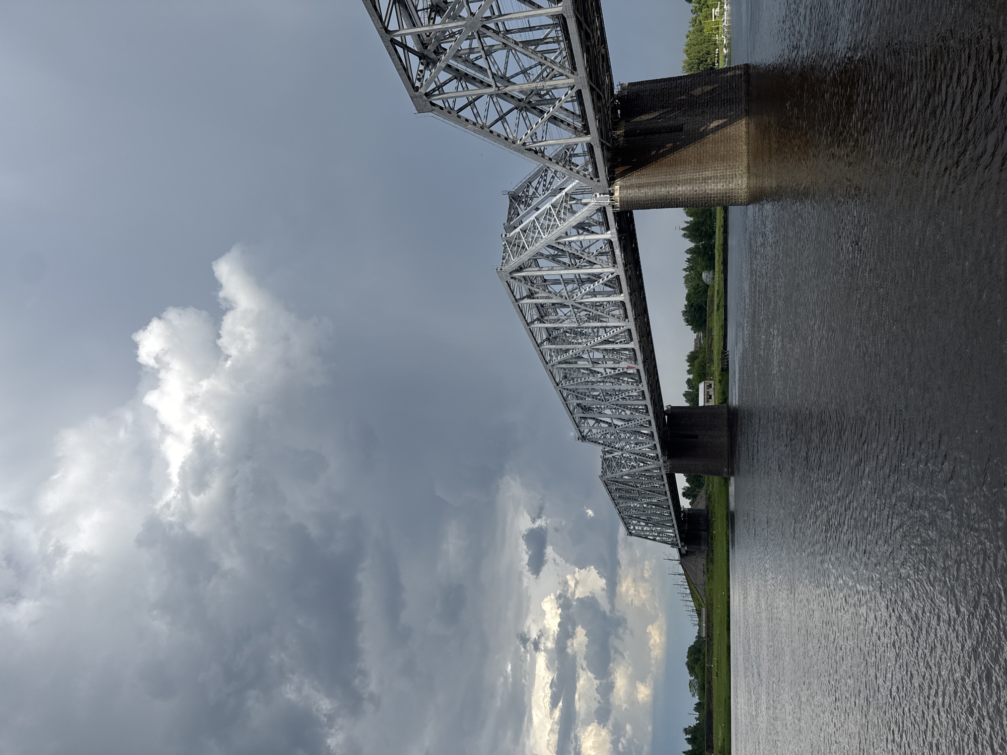 The image shows a railway bridge over a river. The bridge is made of metal truss structures and is supported by stone piers. The river is wide and dark, with ripples on the surface. The sky is cloudy and grey, with some patches of sunlight breaking through. In the background, there are trees and some buildings. The scene is likely in Rusanovo, Russia, given the presence of a railway bridge and the typical landscape. There are no people visible in the image. The environment appears to be a natural setting with man-made structures.