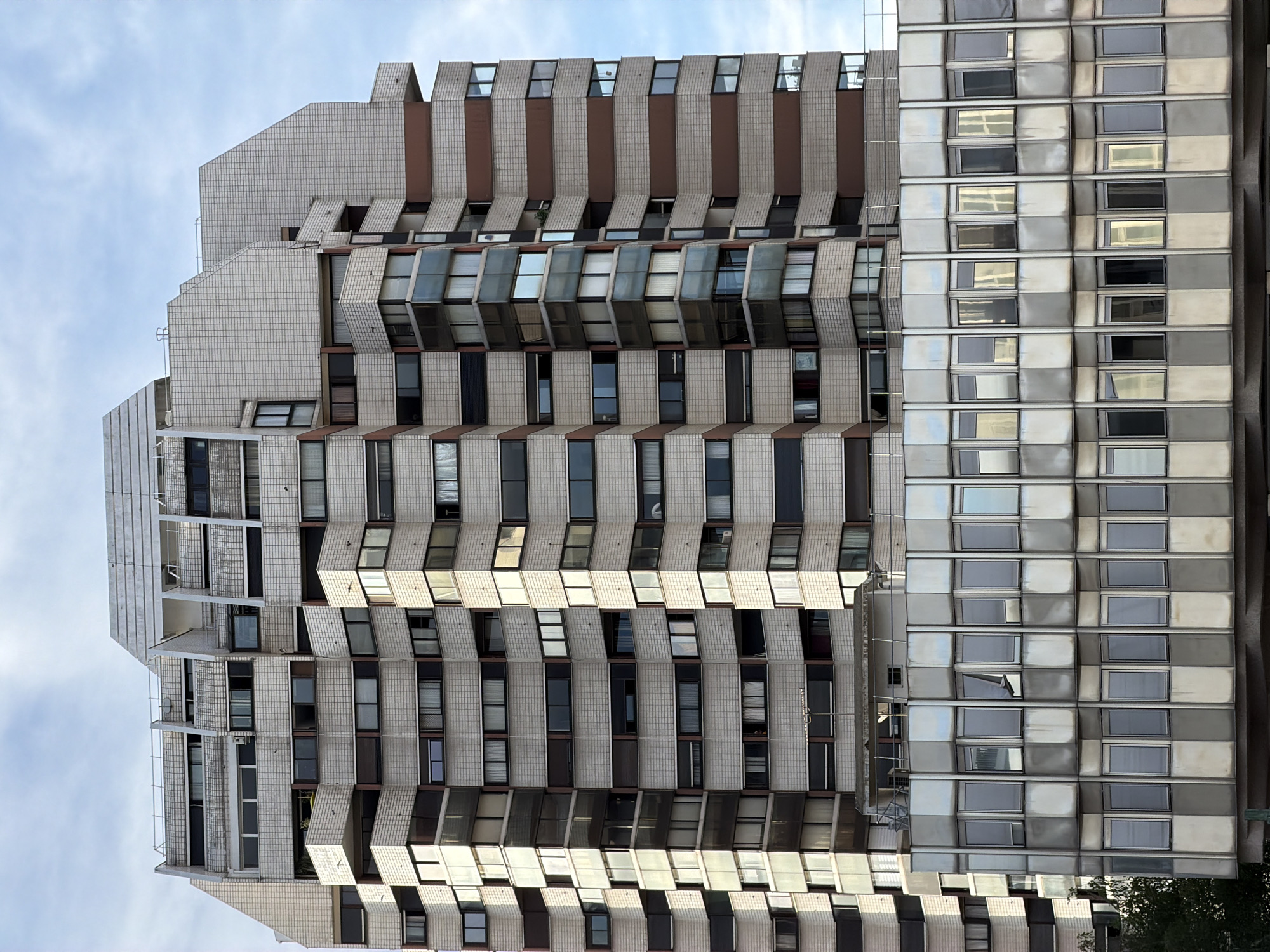 This is a medium-shot, exterior, eye-level, vertical photograph of a Brutalist-style apartment building in Créteil, France. The building is tall and has a stepped, angular facade. The exterior is clad in white tiles and accented with brown and grey panels. There are numerous windows arranged in horizontal rows, some of which are open, revealing darker interiors. The building appears to be uninhabited or at least devoid of visible occupants. To the right of the tiled facade is a section of the building covered in metallic, reflective panels, which mirror the sky and the adjacent facade. The sky is overcast with some patches of blue. The time of day appears to be daytime, likely mid-morning or afternoon, given the overcast lighting. The location is identified as Créteil, France. There is no visible text in the image. The scene evokes a sense of urban density and a somewhat austere architectural style.