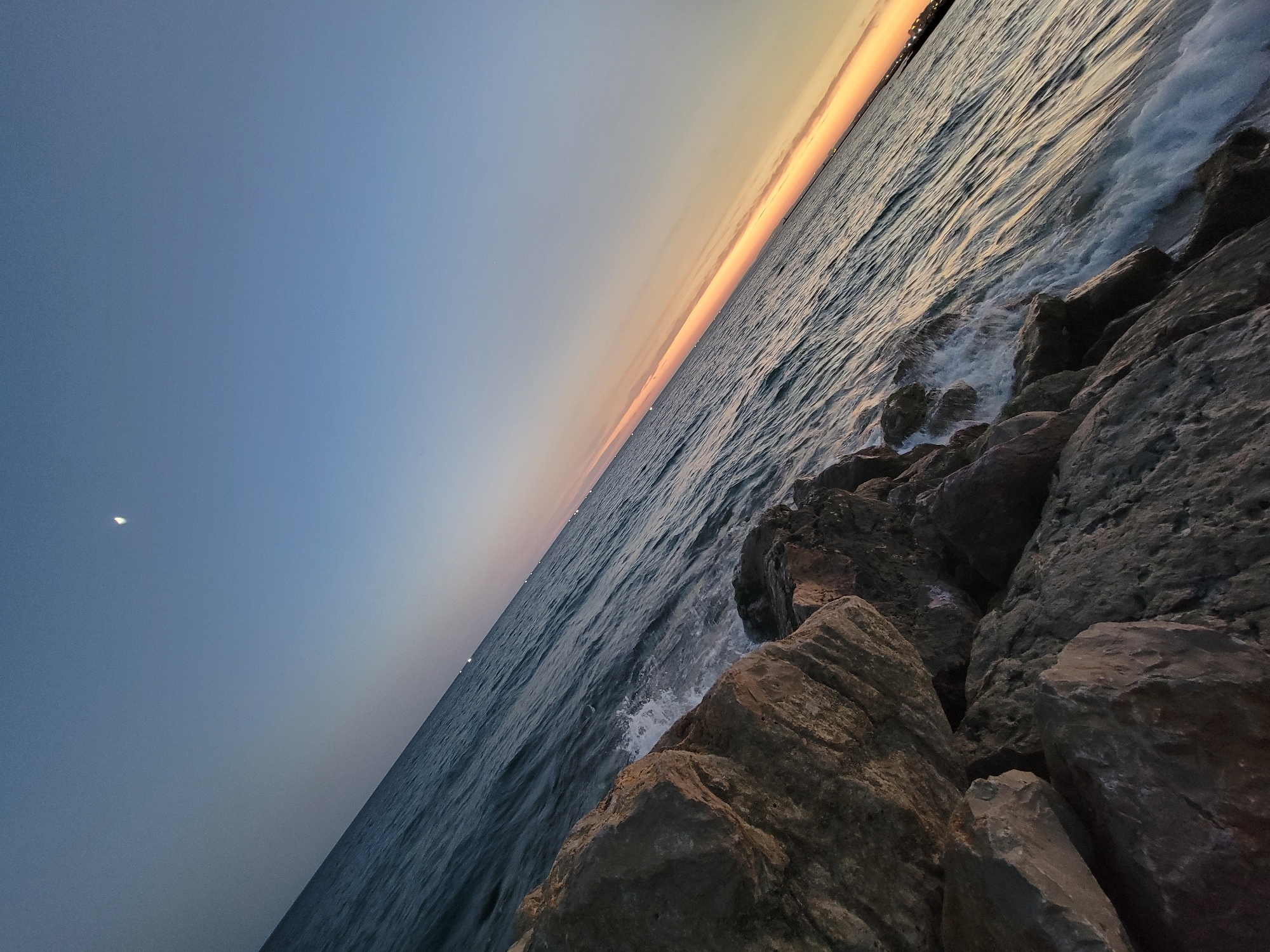 The image captures a diagonal view of the Quarteira coastline at dusk. Large, rough-hewn rocks dominate the foreground on the left, with waves crashing against them. The ocean stretches diagonally across the frame towards the right, its surface rippled. In the distance, a faint band of orange and yellow hues marks the sunset along the horizon. The sky above transitions from a pale blue near the horizon to a deeper blue overhead, with a crescent moon visible in the upper left quadrant. Scattered lights dot the distant coastline, suggesting the presence of urban development. There are no people or animals visible in the frame.