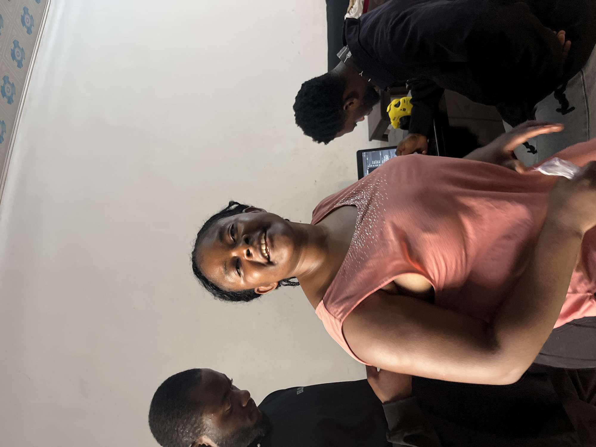 The image depicts an indoor scene, likely a living room, in Gura Top, Nigeria. In the foreground, a woman with dark skin and braided hair is smiling at the camera, wearing a pink sleeveless top adorned with rhinestones. To her left, a man with dark skin, a beard, and short hair is wearing a black sweatshirt and appears to be adjusting something on the woman's left arm. In the background, another man with dark skin and short hair is seated at a table, looking down at a laptop. A bright yellow object, possibly a toy or gadget, is also on the table. The room has a plain white wall and a patterned ceiling. The overall atmosphere appears casual and domestic.