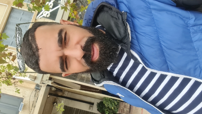 A bearded man with dark hair, wearing sunglasses perched on his head, is shown from the chest up. He has on a blue puffy jacket over a white and navy striped shirt. The background is blurred but appears to be an outdoor setting with building facades and foliage. The image appears to be a casual selfie-style photograph taken at a slightly skewed angle.