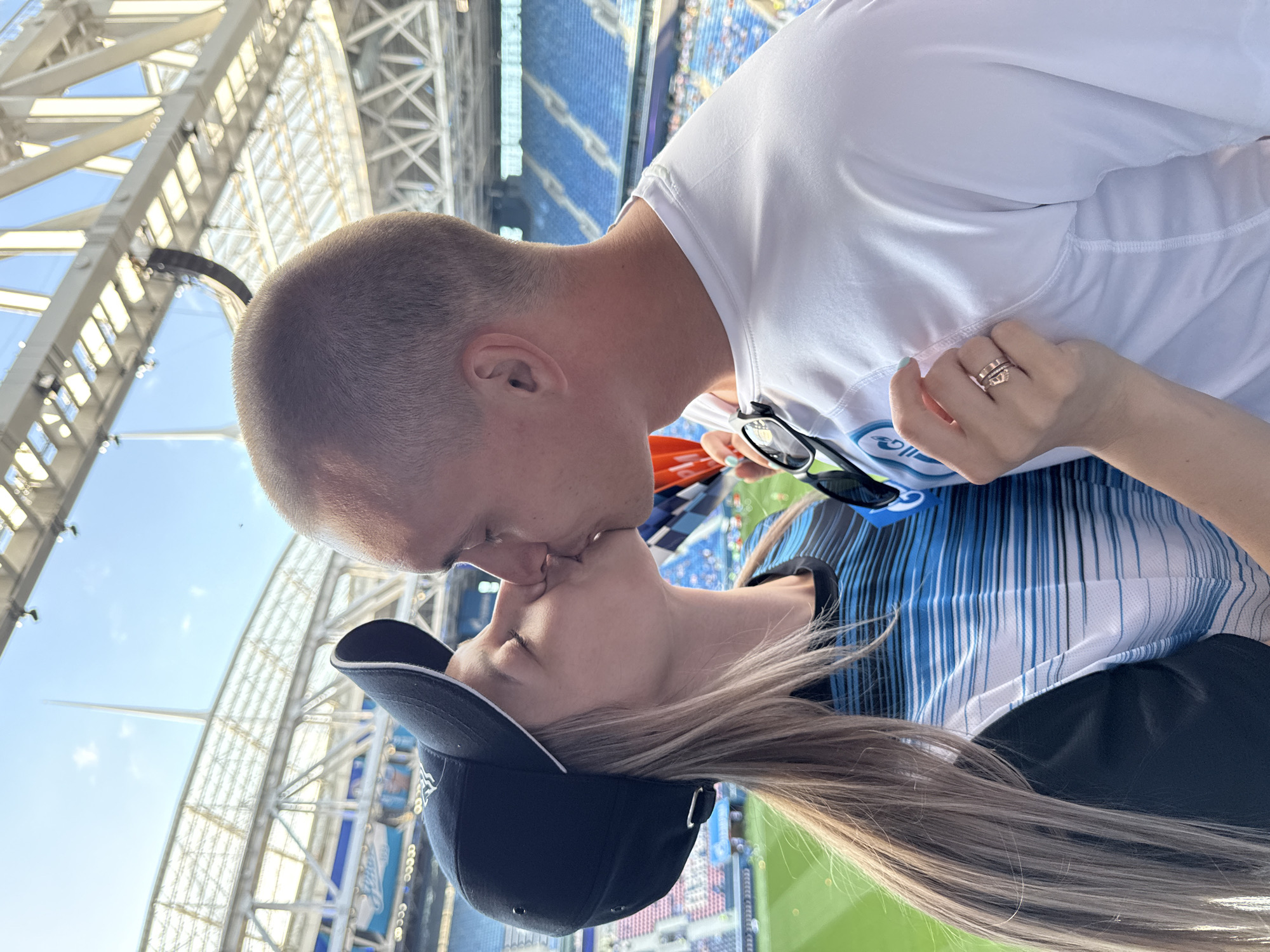 A man and a woman are kissing in front of a stadium. The woman is wearing a black baseball cap and a blue and white striped shirt. The man is wearing a white t-shirt. The stadium appears to be Gazprom Arena, located in Saint Petersburg, Russia. The arena is visible in the background, with blue seats and a green field. The sky is blue and there are some clouds.
