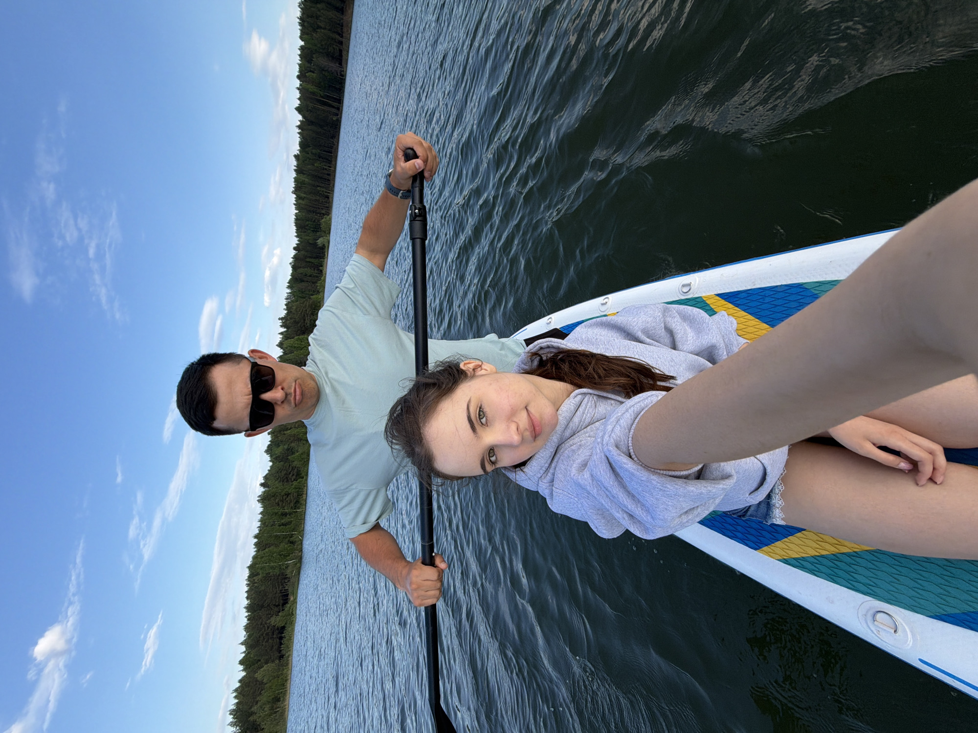 A man and a woman are on a paddleboard in a body of water. The man is holding a paddle and wearing sunglasses and a light green t-shirt. The woman is wearing a light gray hoodie and shorts. The water is dark and wavy. Trees are visible on the shoreline in the distance, and the sky is blue with scattered clouds. The image is taken from a high angle, possibly as a selfie. The location is likely in or near Selychka, Russia, given the prompt context, although this cannot be verified from the image alone.