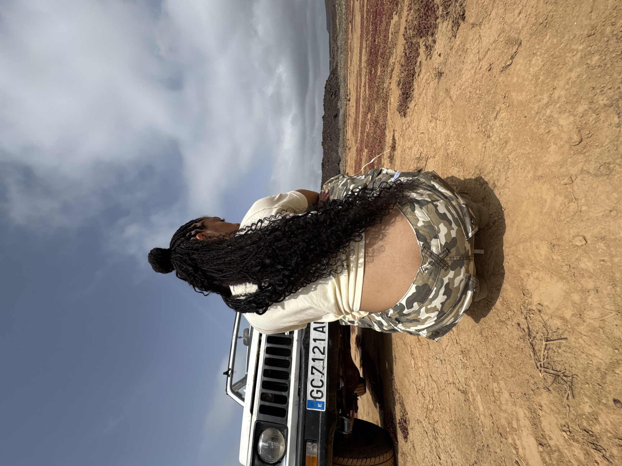 A medium shot depicts a person with long black braids leaning against a white SUV with license plate GC-7121AI in a desert-like area, likely near Tinajo, Spain. The person is wearing a light-colored top and camouflage pants. The landscape includes a rocky, arid ground with sparse vegetation, a range of hills or small mountains in the background, and a partly cloudy sky.