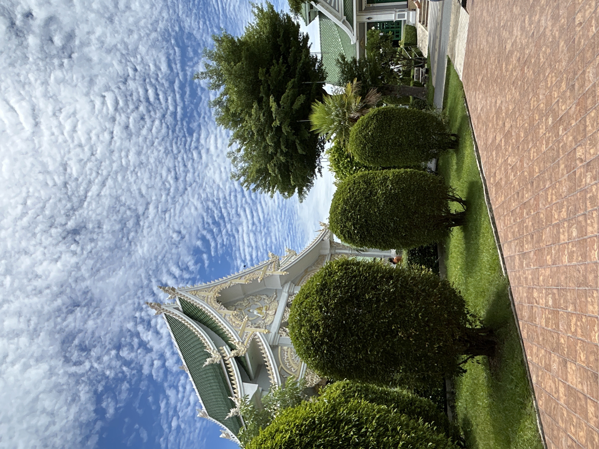 The image depicts a scene in the Thai Ko Subdistrict, Thailand, featuring a traditional Thai temple or building with an ornate green roof and intricate white and gold decorative elements. The architecture is detailed, with pointed roof gables and ornate carvings. In the foreground, there is a meticulously manicured garden with neatly trimmed spherical bushes and a lush green lawn. A reddish-brown tiled pathway runs diagonally across the bottom of the frame. Behind the bushes, a large, mature tree with a full canopy stands prominently. Further back, a portion of another building with a green roof is visible, along with some stairs and a path leading to it. The sky above is a vibrant blue, populated with numerous small, fluffy white clouds arranged in a rippled pattern. The overall impression is one of serenity and traditional aesthetics in a well-maintained outdoor setting.