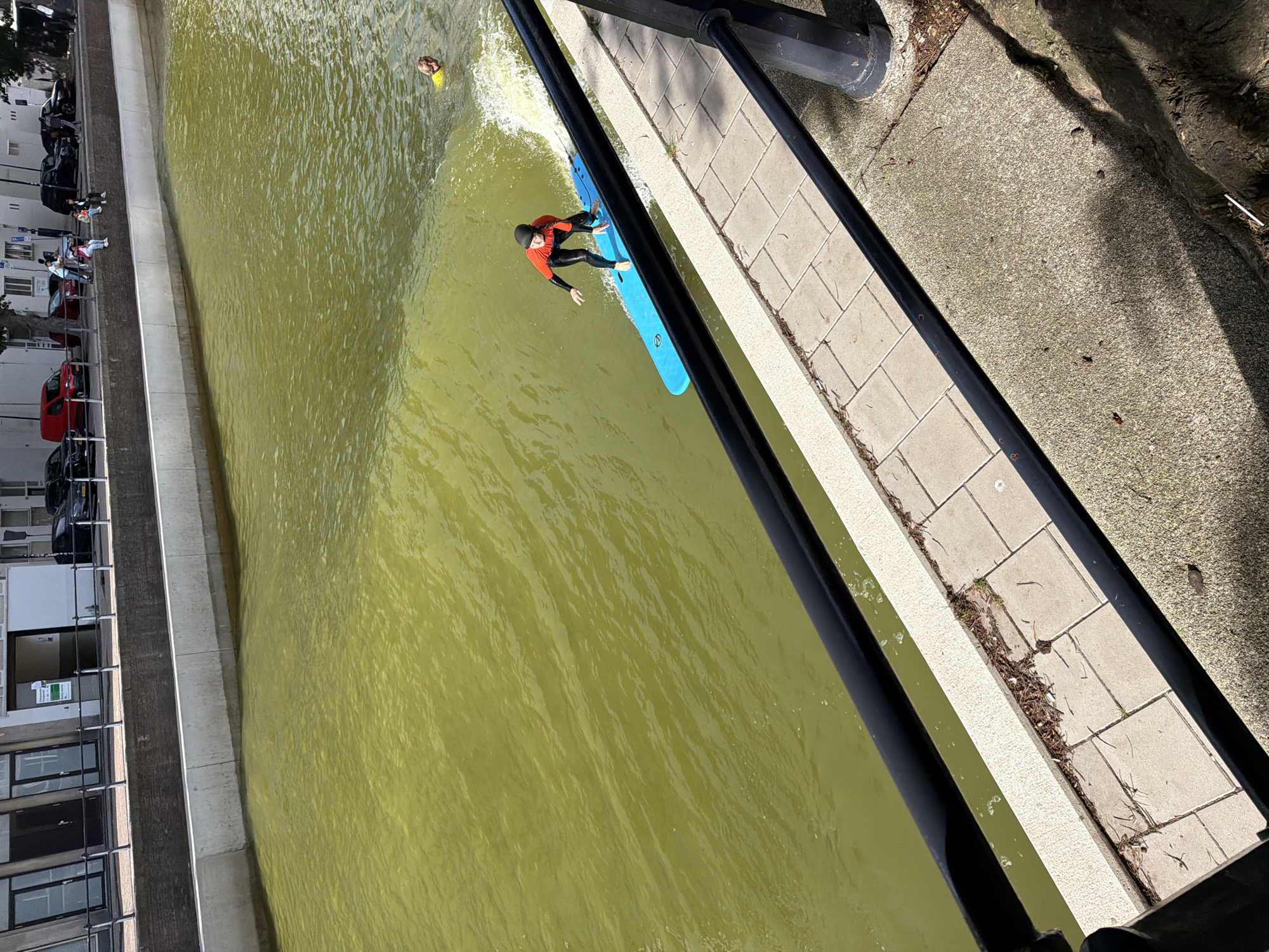 The image depicts a person on a blue surfboard riding a wave in a canal. The person is wearing a red and black wetsuit and a black cap. To the right and further back in the canal, another person in a yellow shirt is visible in the water. The canal is lined with a concrete wall and a paved walkway with a black metal railing. Buildings and parked cars are visible across the canal on the opposite bank. The scene appears to be outdoors on a sunny day. The overall impression is that of a recreational activity taking place in an urban waterway. Based on the architectural style of the buildings and the presence of bicycles and cars, the location is likely a European city.