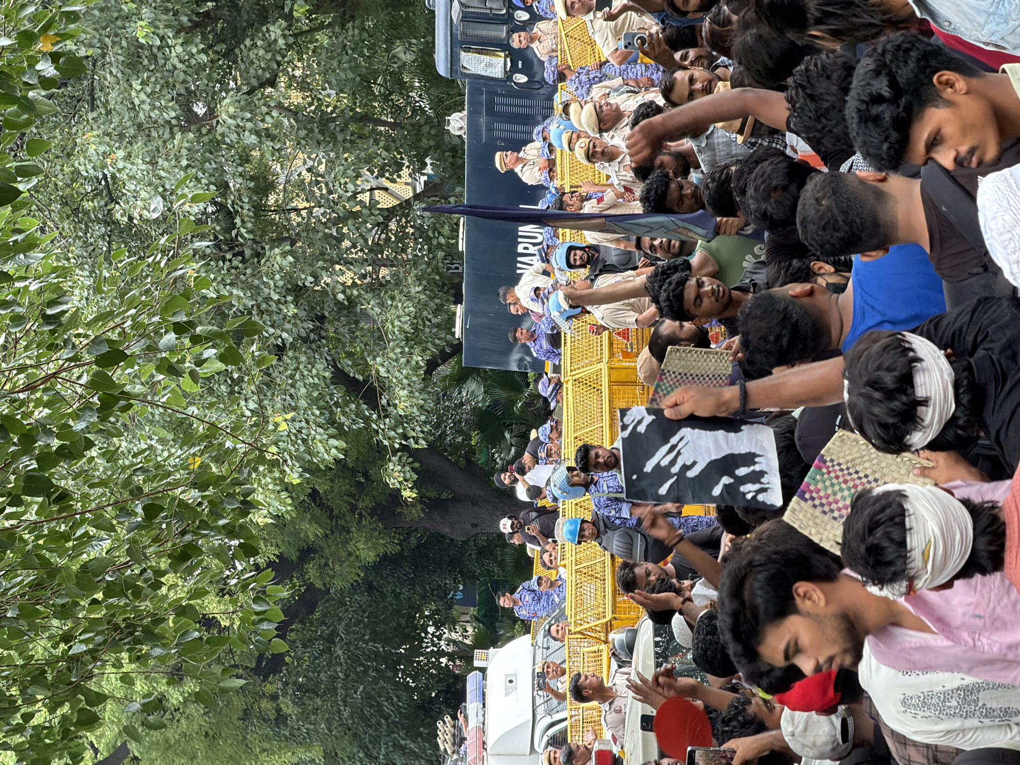 The image depicts a crowd of people, some holding signs and flags, in an outdoor setting. Several individuals in uniform, appearing to be police officers, are visible in the background, some behind yellow barricades. There are also police vehicles present. The environment includes lush green trees overhead, suggesting a park or tree-lined street. One sign held by a protester displays a black and white graphic resembling a handprint. Another individual is holding a blue flag. The overall scene appears to be a protest or demonstration. The presence of police vehicles and officers suggests a security presence at the event. The setting is consistent with an urban environment in New Delhi, India, possibly a public space such as a park or a street near government buildings, where such gatherings often take place.