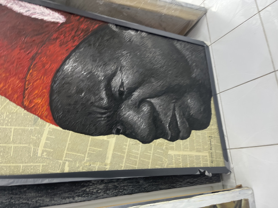 This image captures a framed artwork resting on a white tiled floor in an indoor setting within Alaguntan, Nigeria. The artwork, a textured painting or mixed-media piece, depicts the left profile of a dark-skinned individual, likely an adult male, with a neutral facial expression. The figure wears a prominent red cap, textured with visible brushstrokes, which includes a subtle white feather-like embellishment on its right side. The background of the artwork consists of a collage of light-toned, aged paper sections, appearing as columns of printed text. An inscription, "Adegoke Nurudeen. M 4", is visible in the lower-left section of the artwork's background. The piece is contained within a dark grey or black frame. Other partially visible framed artworks or canvases are present on both the left and right sides of the main subject, leaning vertically against an unseen surface. The floor beneath consists of white square tiles with dark grey grout lines.