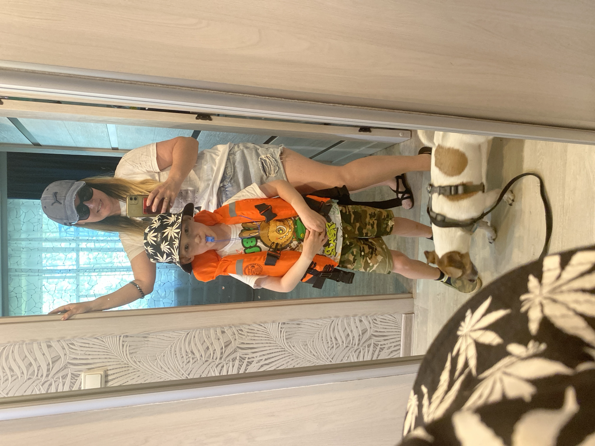 A mirror selfie shows a woman in a gray baseball cap, sunglasses, a white t-shirt, and denim shorts holding a phone, standing next to a young boy wearing an orange life vest and camouflage shorts. The reflection includes a white and brown dog with a harness and leash. The scene takes place indoors, with patterned wallpaper on the lower part of the wall. Noviy, Russia is not depicted.