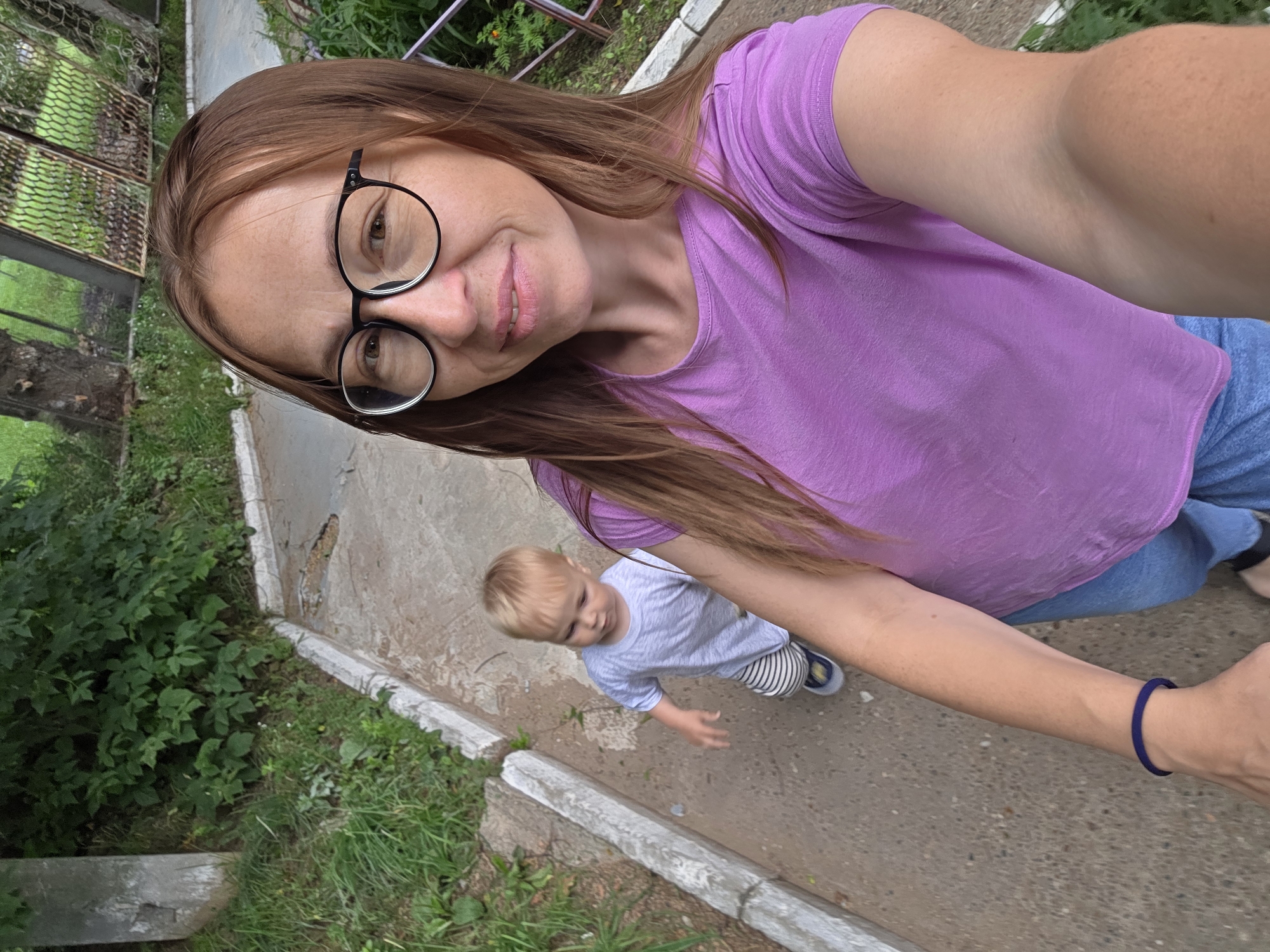 A woman with light brown hair, wearing glasses and a purple t-shirt, takes a selfie outdoors. A young child with blond hair, wearing a grey t-shirt and striped pants, is visible behind her, walking on a paved path. The environment appears to be a park or a residential area, with greenery, trees, and a fence in the background. The image seems to have been taken on a cloudy day. The location is identified as Chaikovsky, Russia.