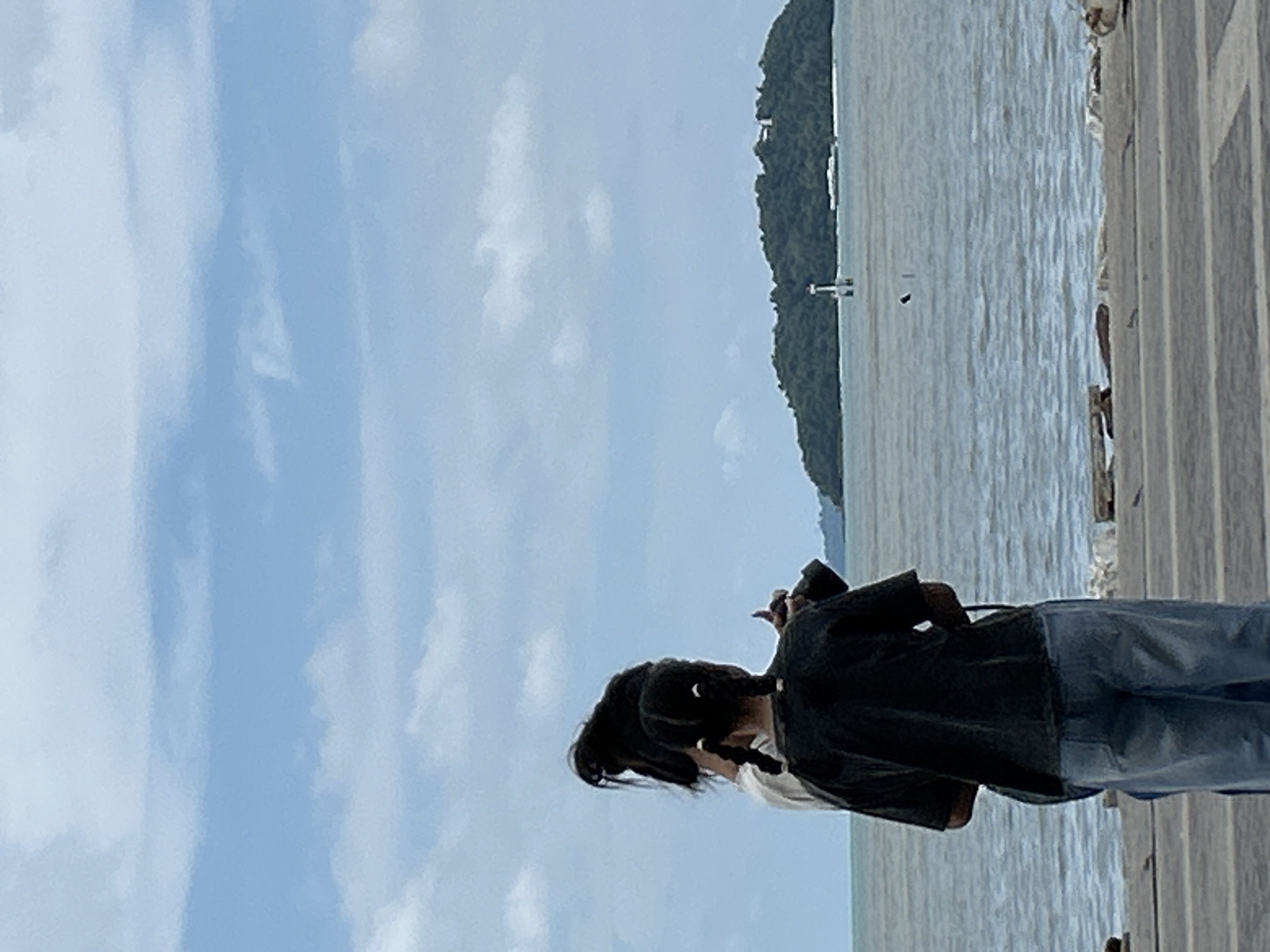 Two individuals are standing on a paved walkway facing a body of water. The person in the foreground is wearing a dark t-shirt and light blue jeans, with their hair styled in two pigtails. They are holding a dark object, possibly a camera, in their hands. Behind them, another individual, whose face is not visible, is wearing a light-colored top. The water extends to the horizon, where a green, tree-covered island is visible with a white structure on its shore. The sky is blue with scattered white clouds. The location is a waterfront area within Phuket City Municipality, Thailand.