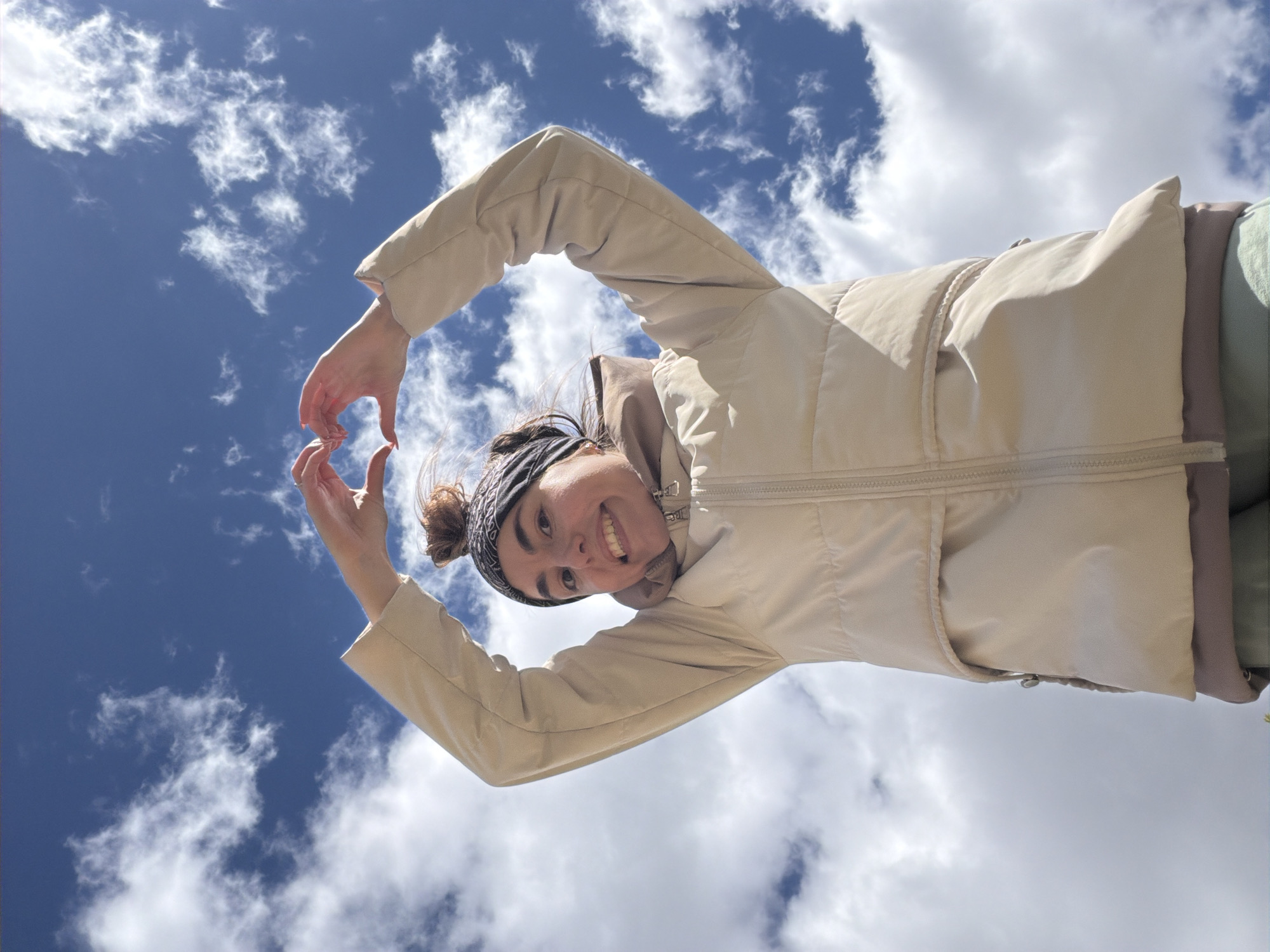 A young woman is centered in the frame, smiling directly at the viewer while making a heart shape with her hands. She is wearing a light beige puffer jacket and a patterned headband. Her hair is styled in a bun. The backdrop is a vivid blue sky filled with fluffy white clouds. The perspective is from below, looking up at the woman, giving the impression that she is standing above the viewer. The image does not contain any information indicating this location is in Nizhnii Vishur, Russia.