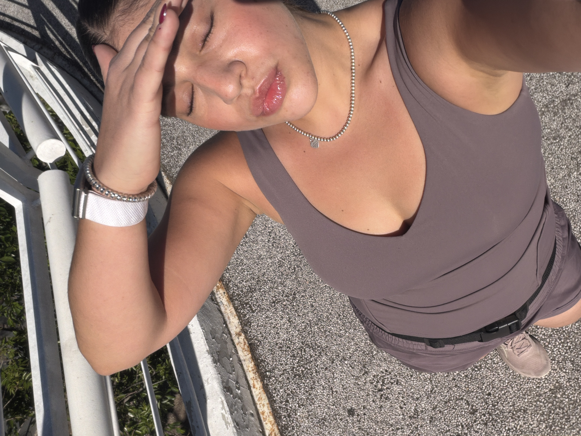 An overhead selfie captures a young woman outdoors in bright daylight, possibly on a balcony or rooftop in Puebla, Mexico. Her right hand is raised, shielding her eyes from the intense sun, causing them to be closed. Her lips are slightly parted and glossy.
She is dressed in a muted purple or taupe athletic tank top and matching shorts, cinched with a black belt that has a simple black buckle. Accessories include a silver beaded necklace with a small circular pendant resting on her chest, and on her left wrist, a silver beaded bracelet along with a white smartwatch featuring a textured strap. Light-colored, possibly beige or taupe, sneakers are visible on her right foot. Her fingernails are painted a dark red or burgundy.
The background features a white metal railing to her left, through which green foliage is visible. The ground is a light grey, pebbled or textured surface, likely concrete. Strong shadows cast by the railing indicate clear, sunny weather. The overall impression is one of casual comfort in bright, warm conditions. No visible text is present in the image.