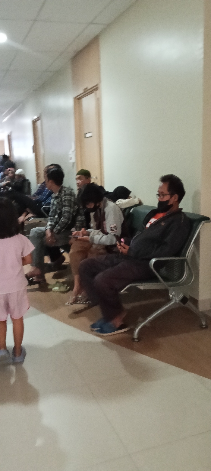 The image shows a waiting area in a hospital or clinic located in Soreang, Indonesia, at coordinates -7.032 latitude, 107.527 longitude. There are several people seated on a long, green-cushioned bench and a single chair, while a young girl stands to the left of the frame. The man on the chair is wearing a black jacket and a face mask, while the person to his left is wearing a white jacket with a black design on the left shoulder. Both are looking down at their phones. The person seated to the left of the man in the white jacket is partially visible, wearing a brown shirt and plaid shirt over it.  Behind the seated people, a woman wearing a black jacket and a face mask is seated with her back turned to the camera.  A man wearing a green hat and a blue shirt is seated to her left. There are several people in the background, but they are obscured.  The walls of the waiting area are light beige, and the tiled floor has a pattern of light beige and dark brown tiles.  The waiting area is well-lit and appears clean.  The scene is one of quiet contemplation and waiting.