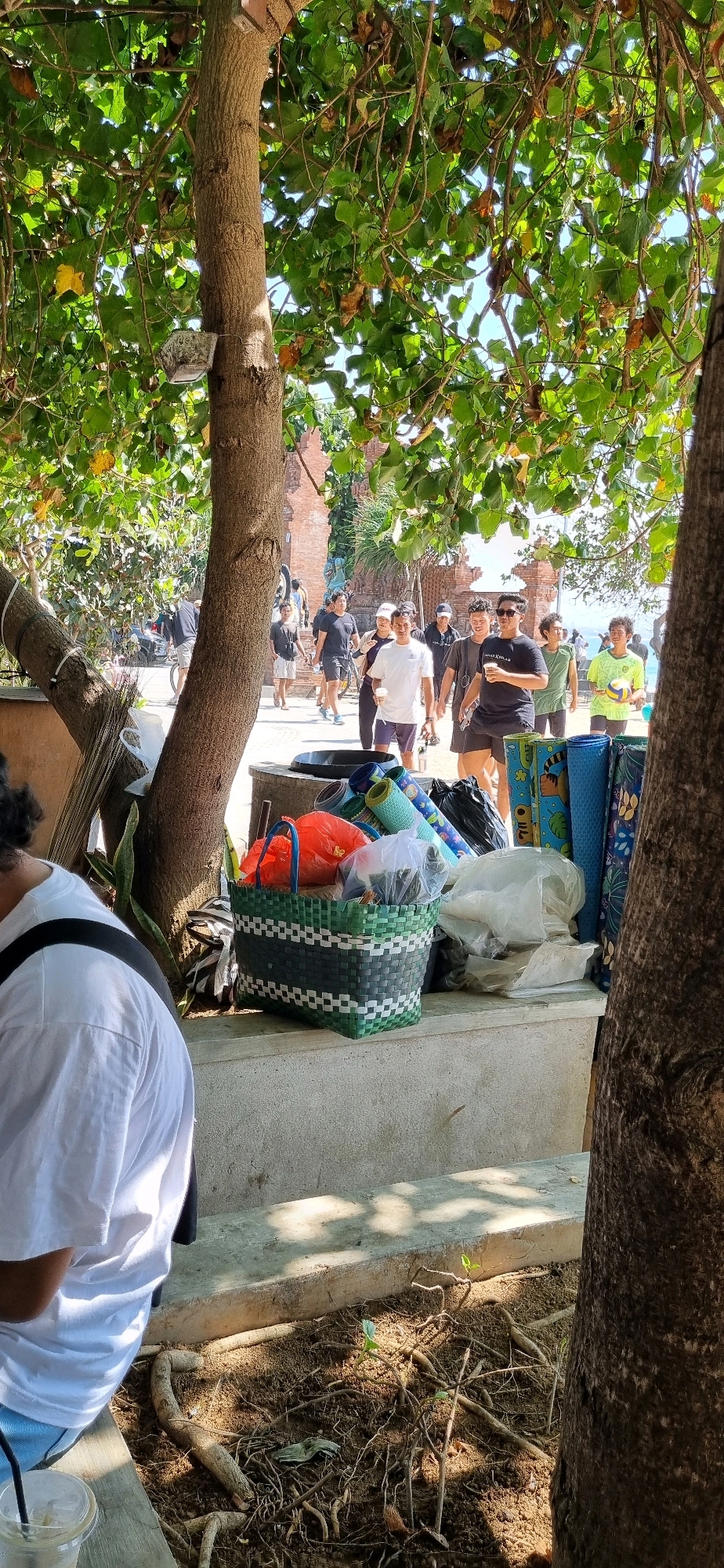 The image depicts a sunny outdoor scene in Denpasar, Indonesia, likely a public area near the coast given the presence of people in casual attire and beach-like items. In the foreground, a person wearing a white t-shirt and blue shorts is partially visible, facing away from the camera. Next to them is a clear plastic cup with a straw.
To the right, a large tree trunk dominates the foreground. In the mid-ground, there is a concrete structure with various items placed on top, including a green and black woven basket filled with orange plastic bags and other assorted items, several rolled-up mats in different colors and patterns, and black trash bags. Behind this, a group of people are walking, some carrying bags and one person holding a soccer ball. They are dressed in casual clothing such as t-shirts and shorts.
The background shows a pathway or road with more people walking and some parked vehicles. There appears to be a brick structure, possibly an archway or part of a building, in the distance. The overall environment suggests a lively, possibly tourist-oriented area with lush green foliage from trees overhead, casting dappled sunlight on the ground and objects. The sky is bright and appears clear.